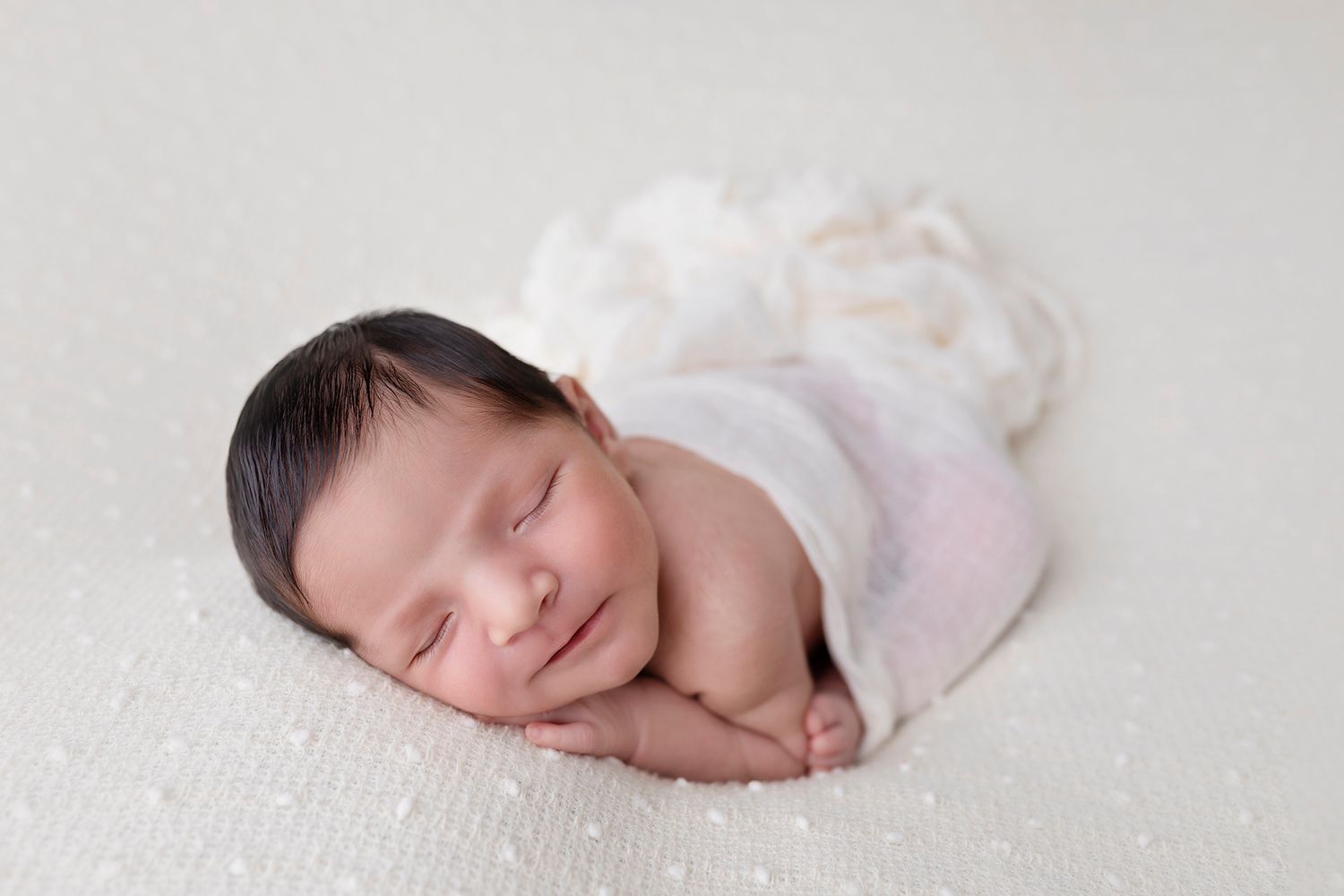 Peaceful sleeping newborn swaddled in white fabric on textured white background.