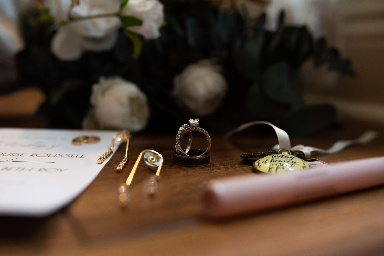Wedding rings and jewelry arranged with flowers and paper on a dark wooden surface.