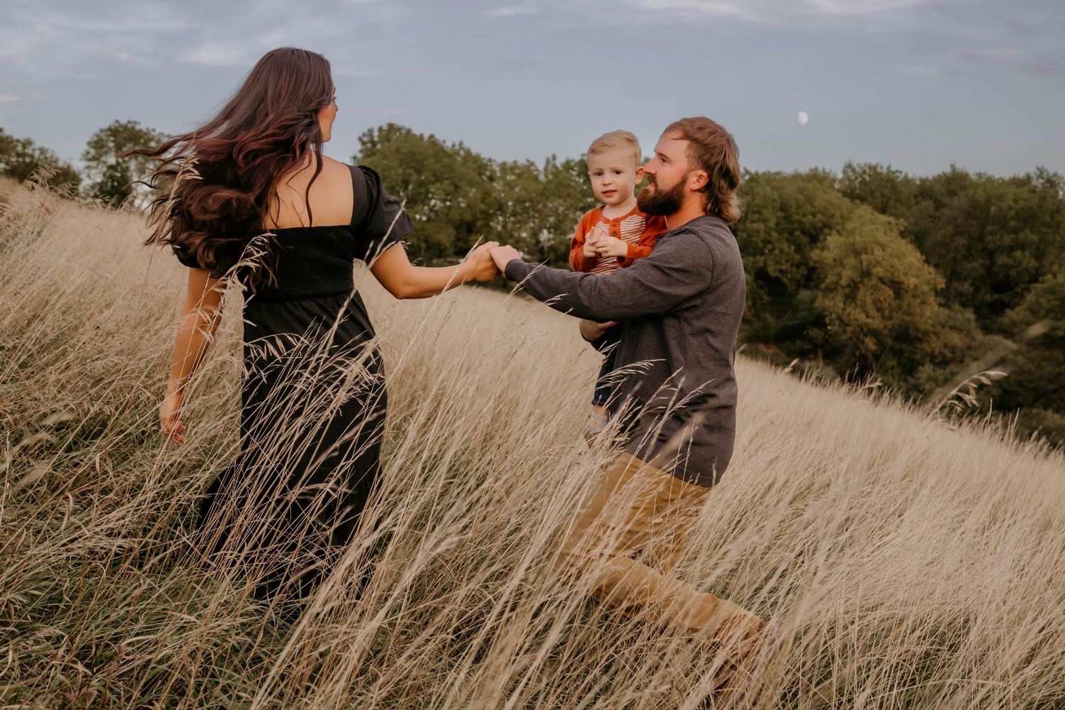 Family of three playing in tall golden grass field, father holding baby while reaching for mothers hand.