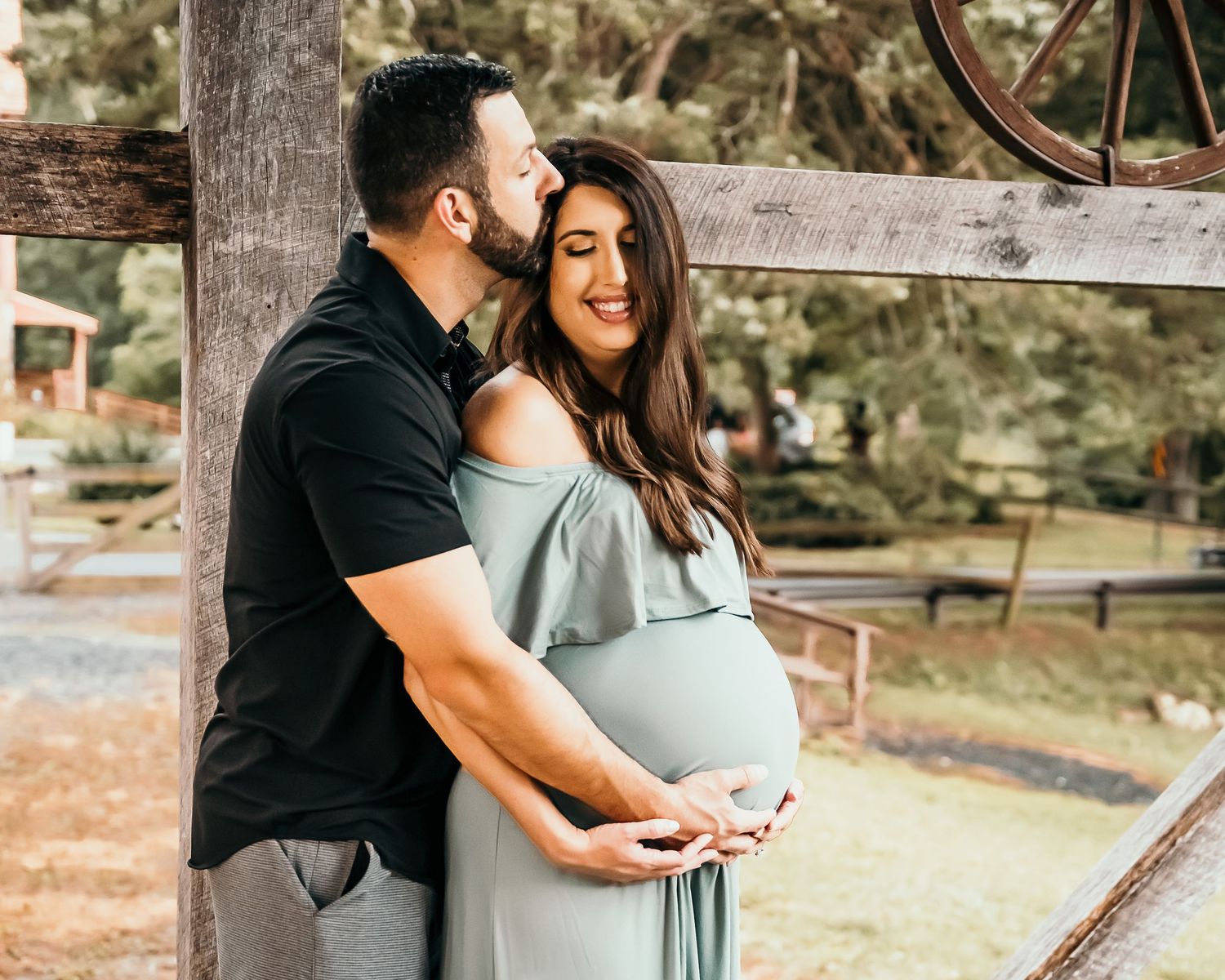 A couple in a black shirt and blue dress embrace outdoors while cradling a pregnant belly in a tender moment.