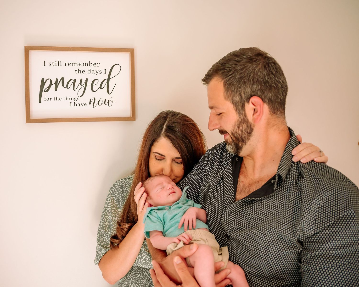 Parents share a tender moment with their newborn baby while standing near a wall with framed artwork.