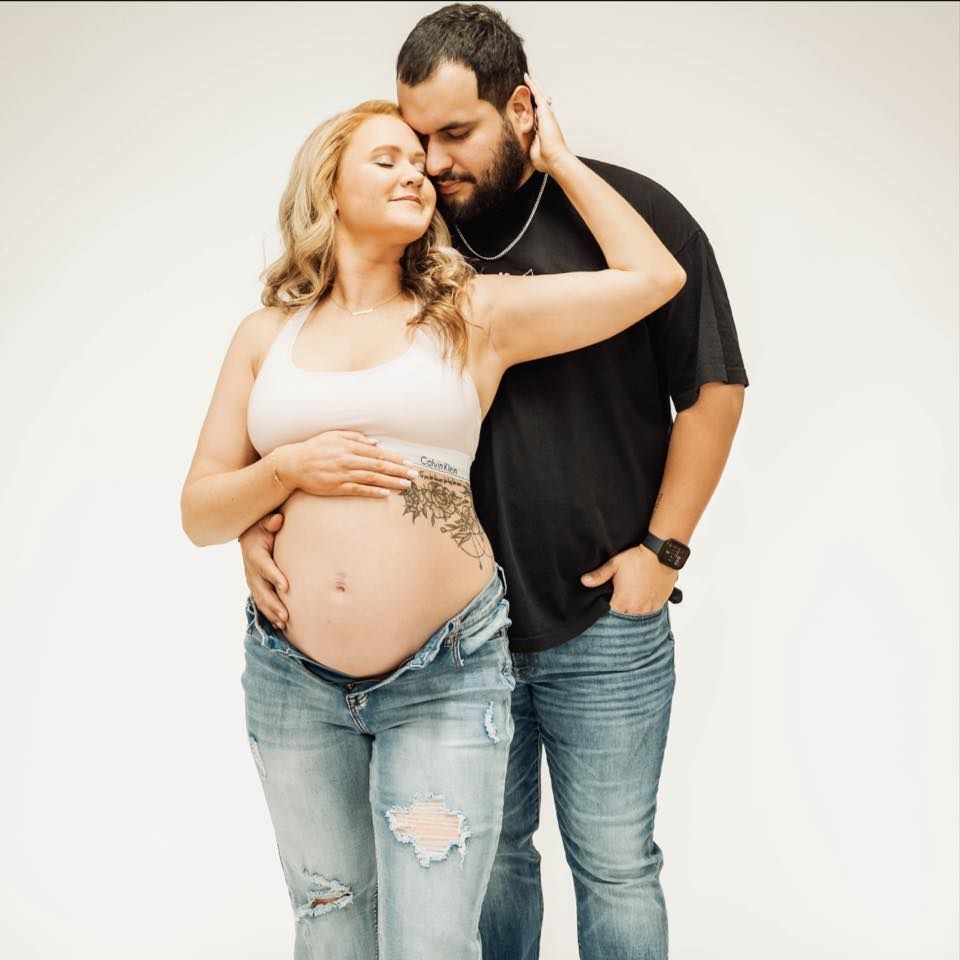 Intimate maternity photo of an expectant couple in casual attire embracing against white background, showing henna belly art.