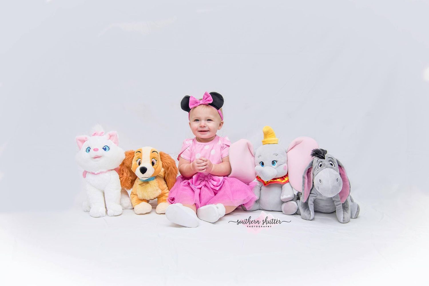 A baby in a pink dress and Minnie Mouse ears sits surrounded by plush stuffed animals on a white background.