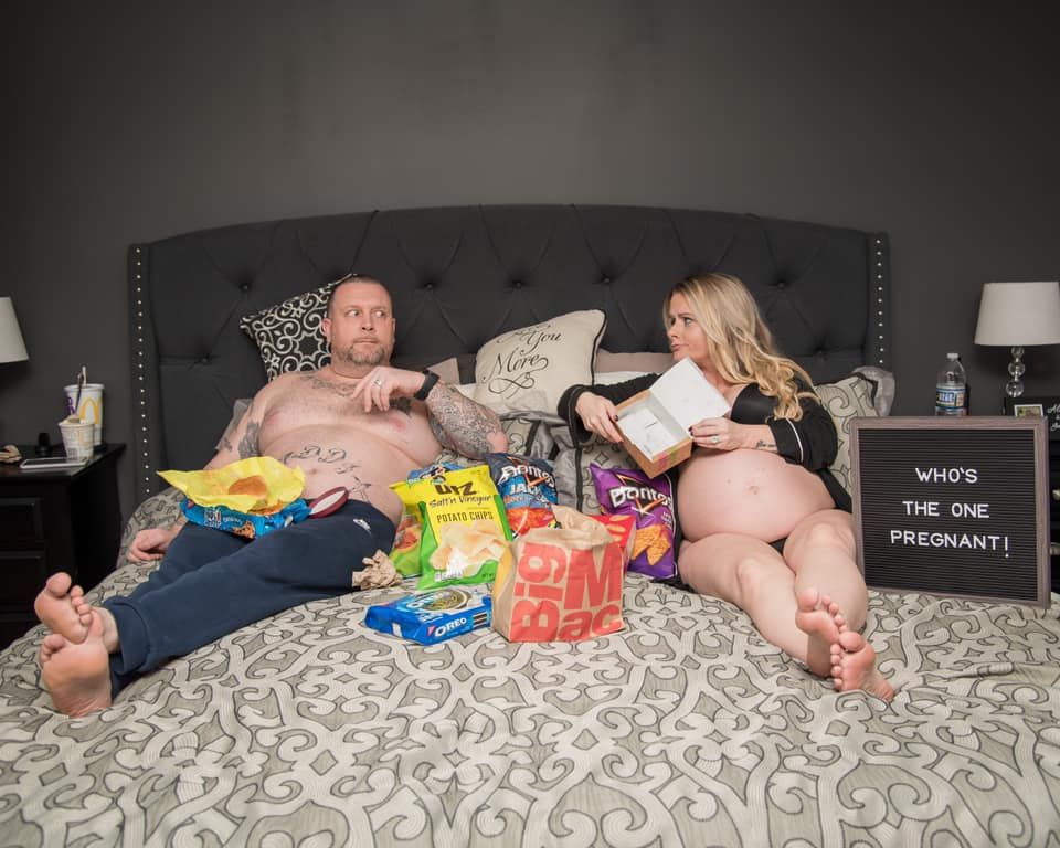 Two people lounging on a bed surrounded by snacks and junk food with a sign that reads "Who's the one pregnant?".