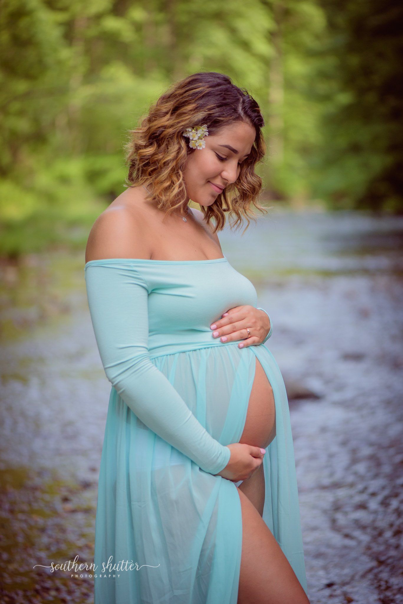 A mother-to-be in a flowing mint maternity gown poses by a stream in an outdoor pregnancy photo session.