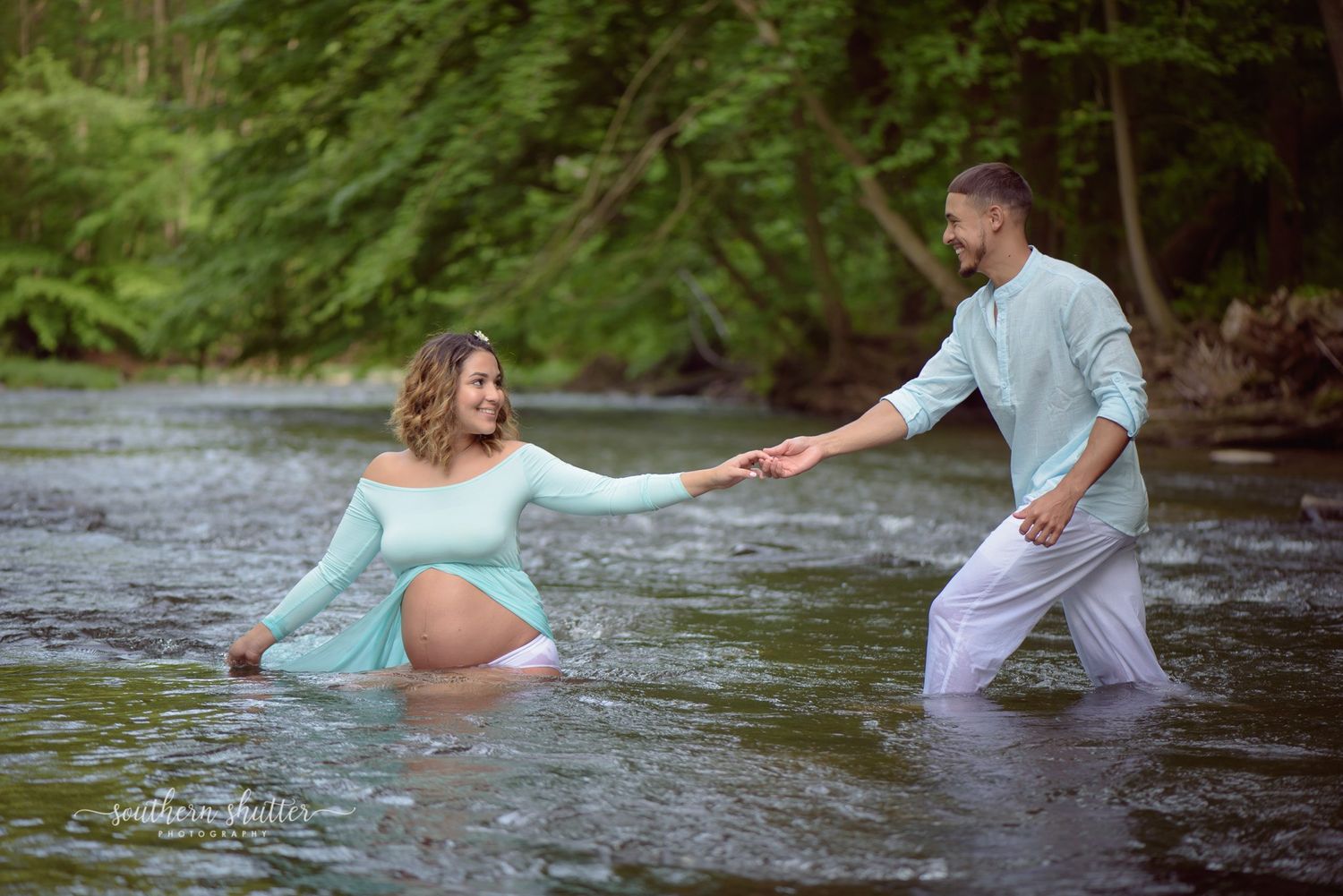 Two people hold hands while standing in a flowing stream with forest trees in the background.  Maternity shoot