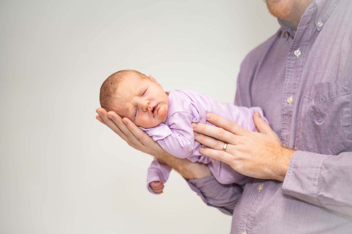 A sleeping newborn baby wearing a lavender onesie is gently cradled in outstretched hands against a light background.