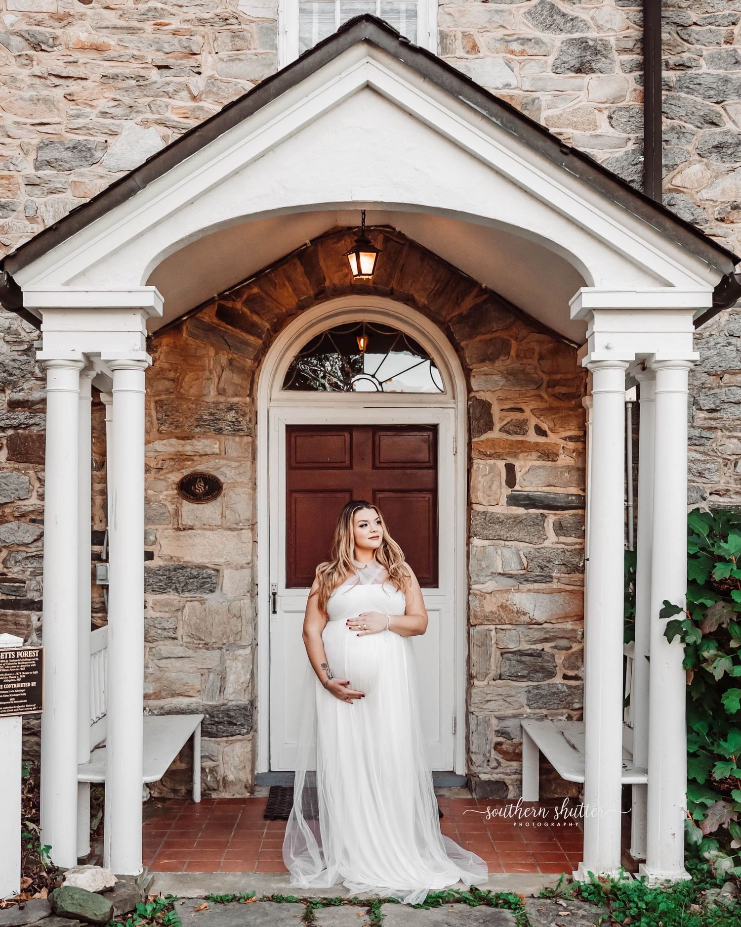 An elegant stone church entrance with white columns frames someone in a flowing white maternity gown.