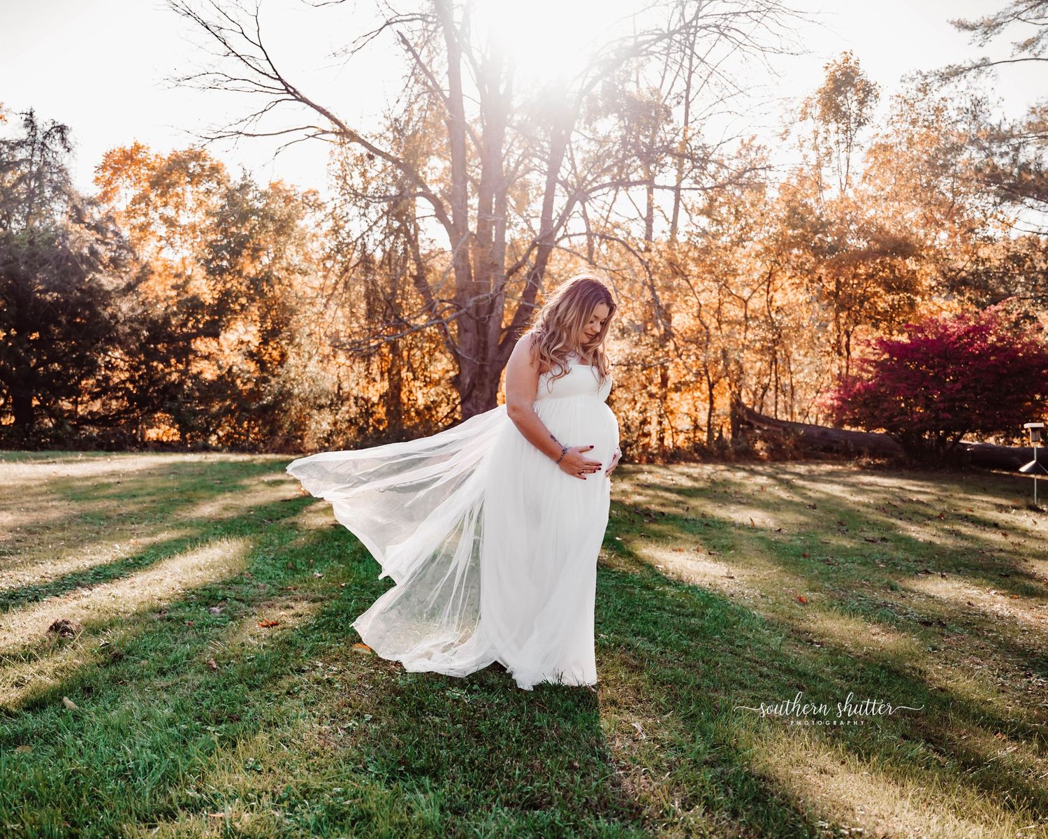 A flowing white maternity dress catches the sunlight in a dreamy autumn forest setting during golden hour.