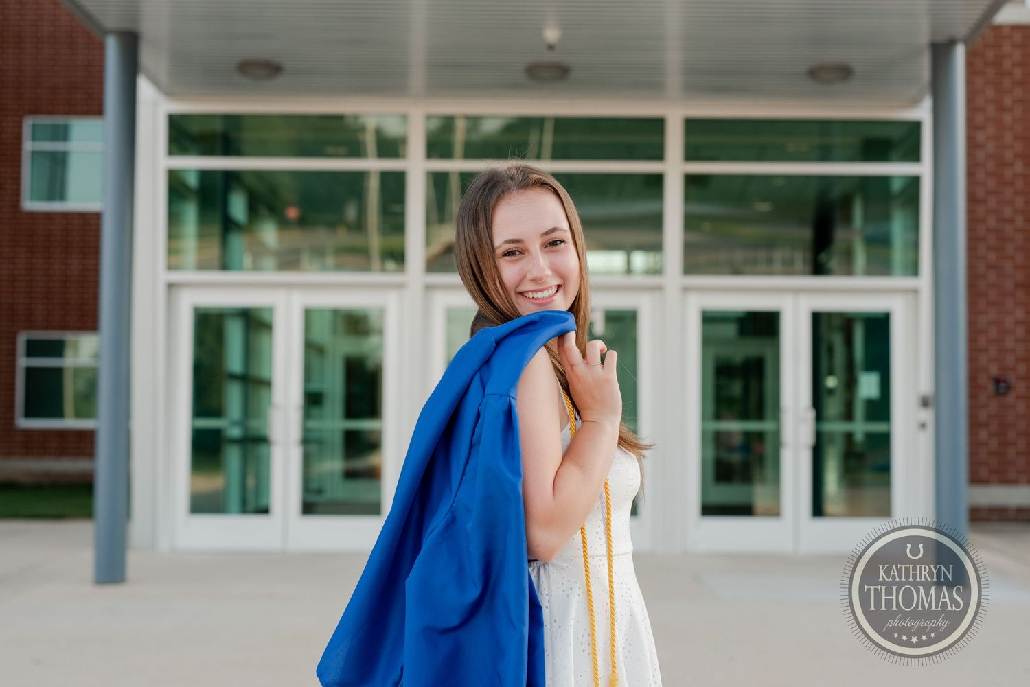 Graduate in a white dress and blue cap and gown poses in front of Spring Grove Area High School entrance.
