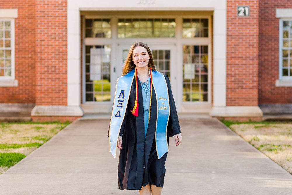 Academic graduation attire with light blue stole and black gown in front of brick building.