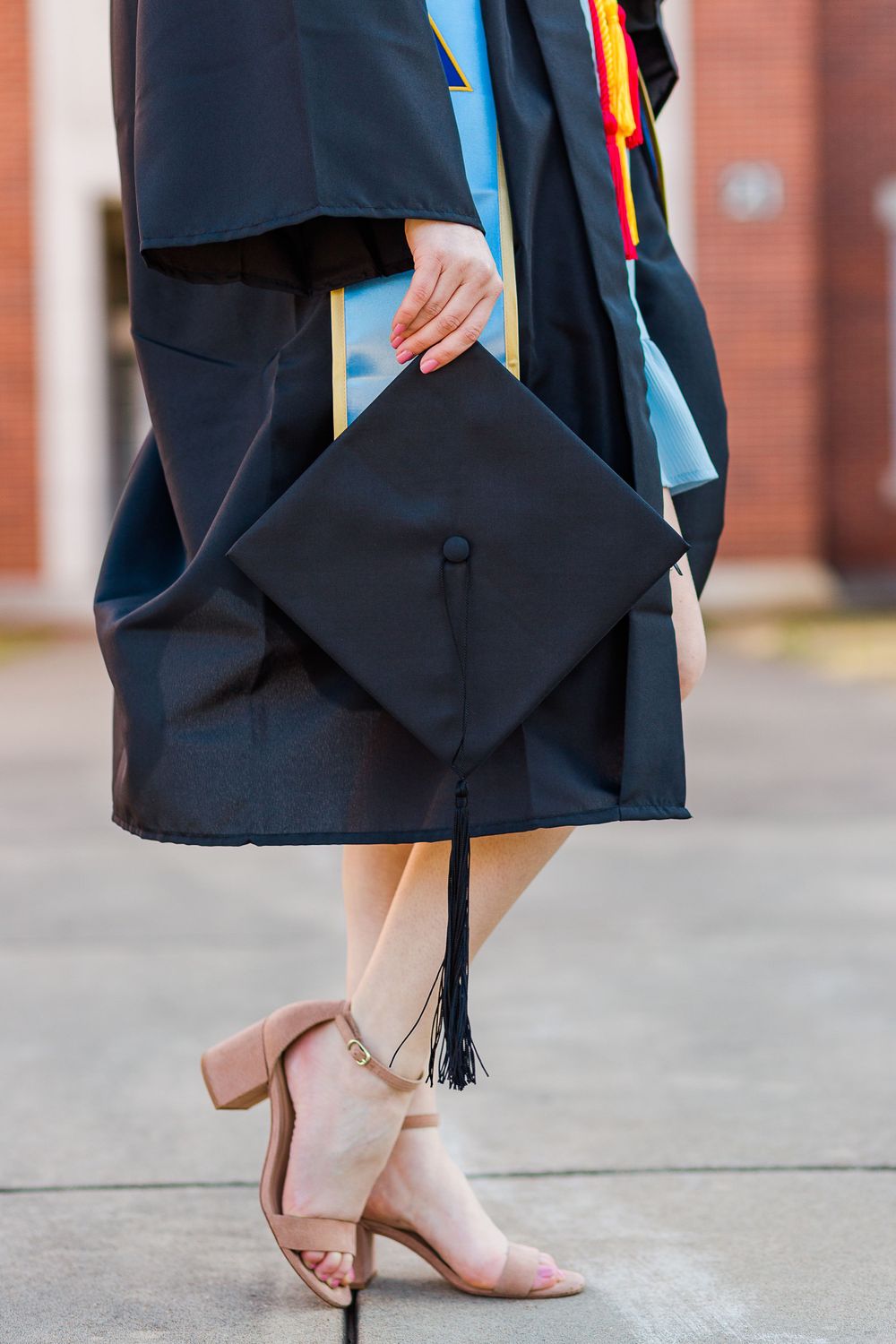 Graduate wearing black cap and gown holds graduation cap while showing off tan heels.