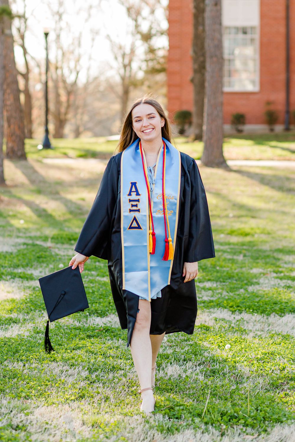 Graduate in academic regalia with decorated stole and cap holds graduation cap on sunny campus lawn.