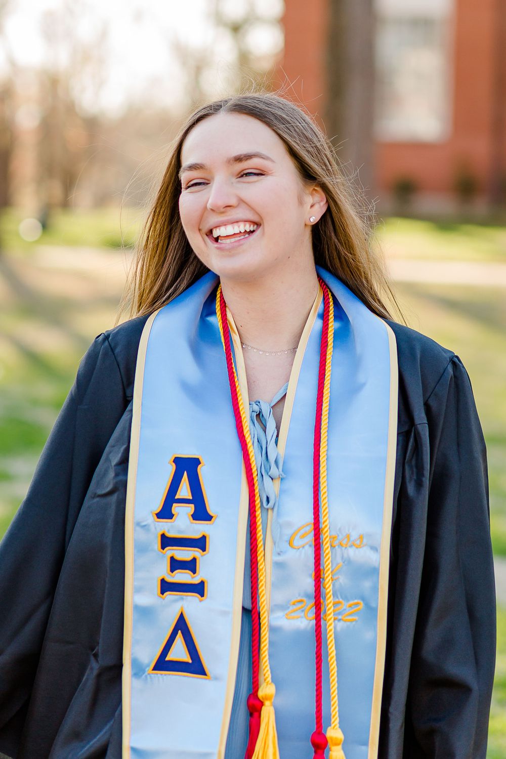 Graduate wearing light blue Alpha Xi Delta sorority stole with colorful cords at outdoor ceremony.