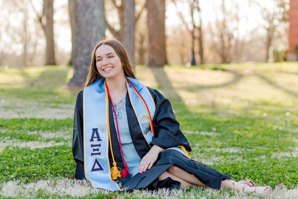 Graduate in black gown and AXiD stole sits on grass for outdoor portrait.