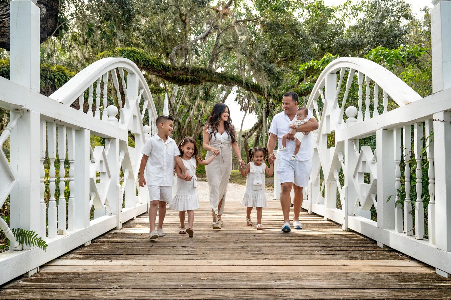 A family in white outfits walking together across a white wooden bridge surrounded by trees.
