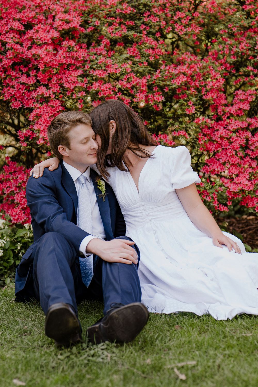 Bride whispering in grooms ear while sitting on ground with red flowers in background