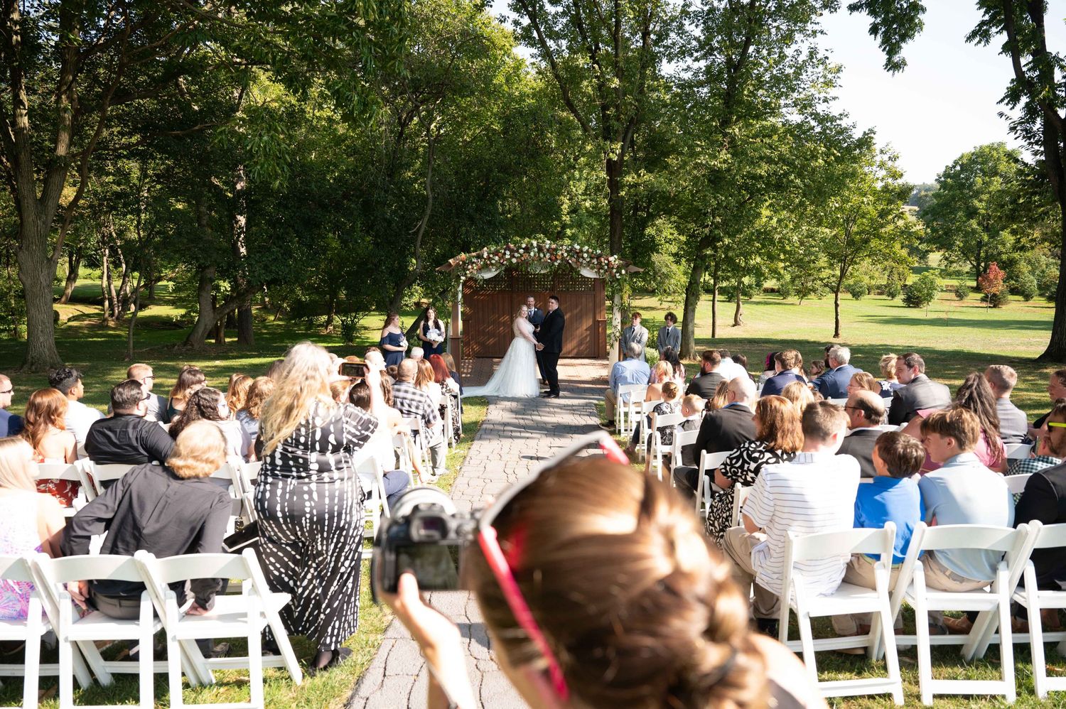 Sun-dappled outdoor wedding ceremony with guests in white chairs facing floral arch.