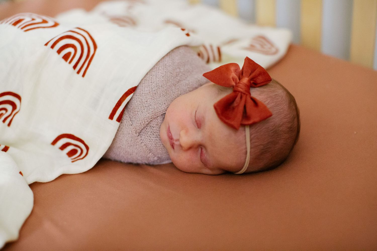 A sleeping newborn baby wearing a red bow headband and wrapped in a rainbow print blanket.