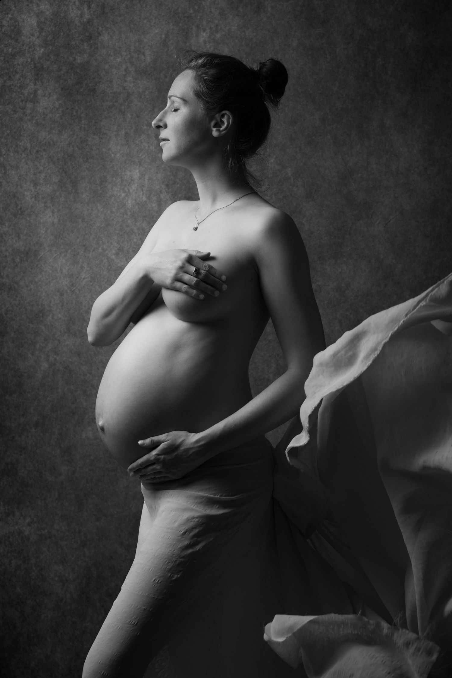 Black and white artistic maternity portrait of pregnant woman with flowing fabric against dark background.