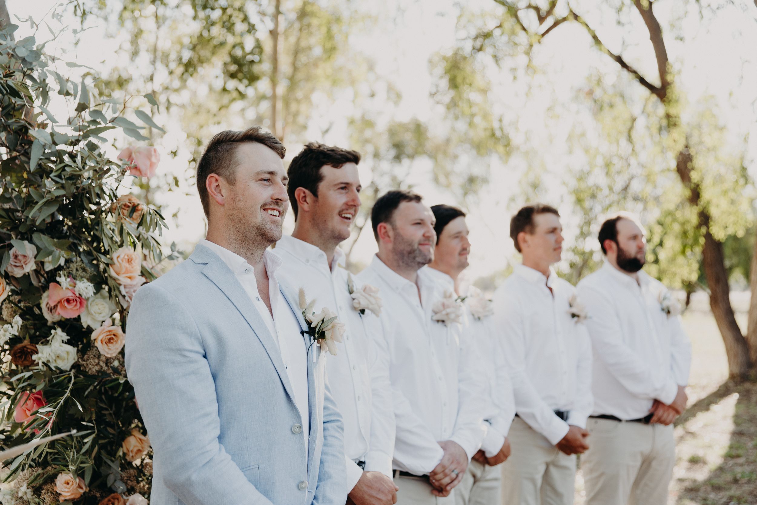 Wedding party members in white attire standing together under trees at an outdoor ceremony.