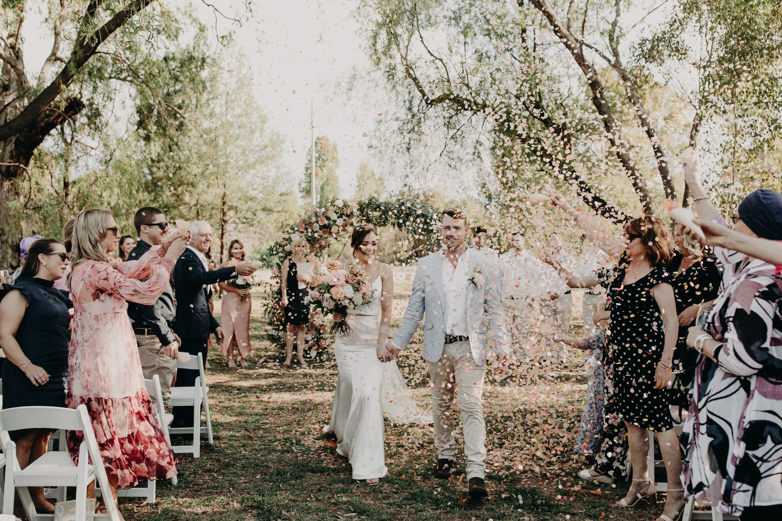 Wedding guests celebrate with confetti toss under trees at outdoor ceremony.