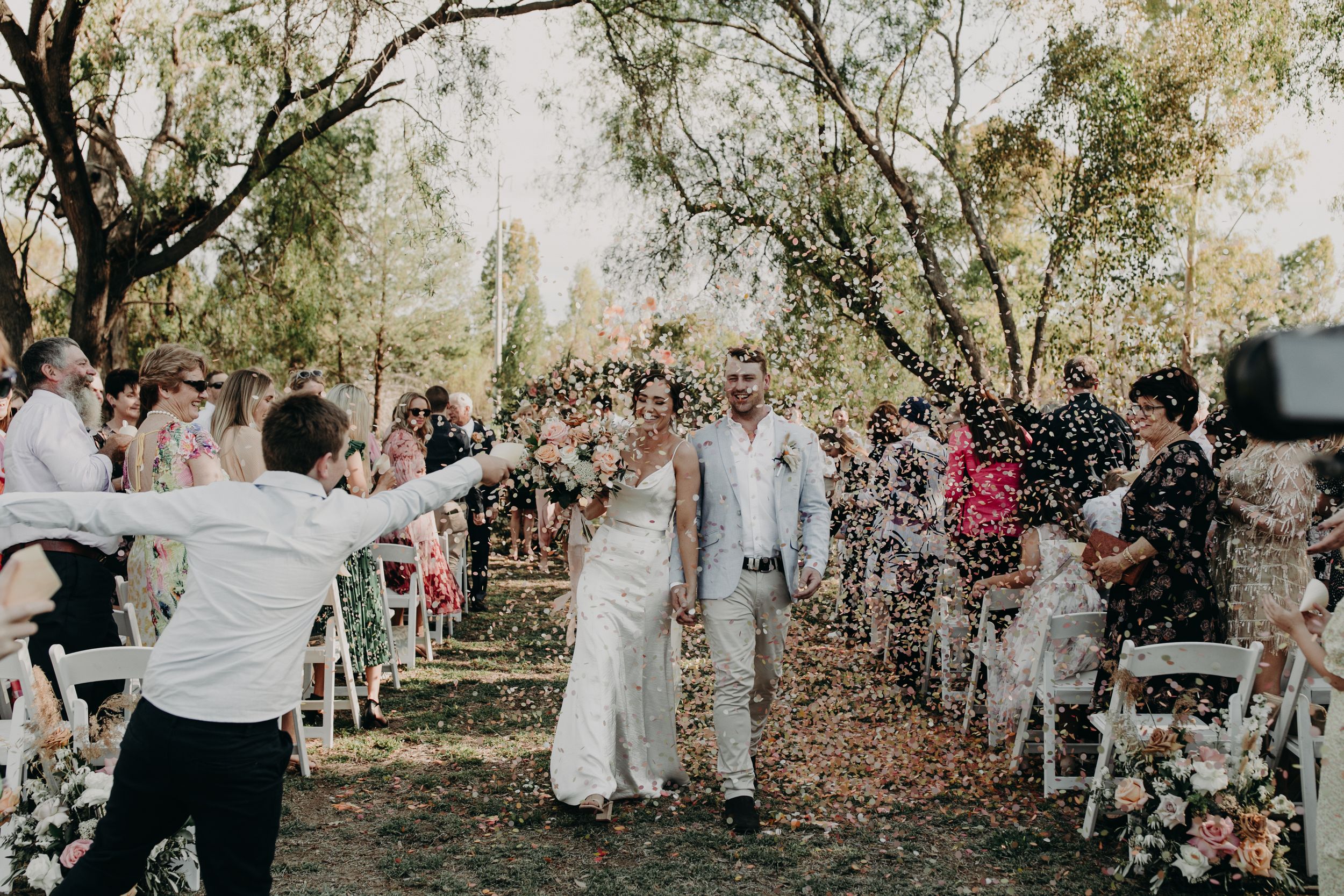 Outdoor wedding celebration with guests throwing confetti in a wooded setting.