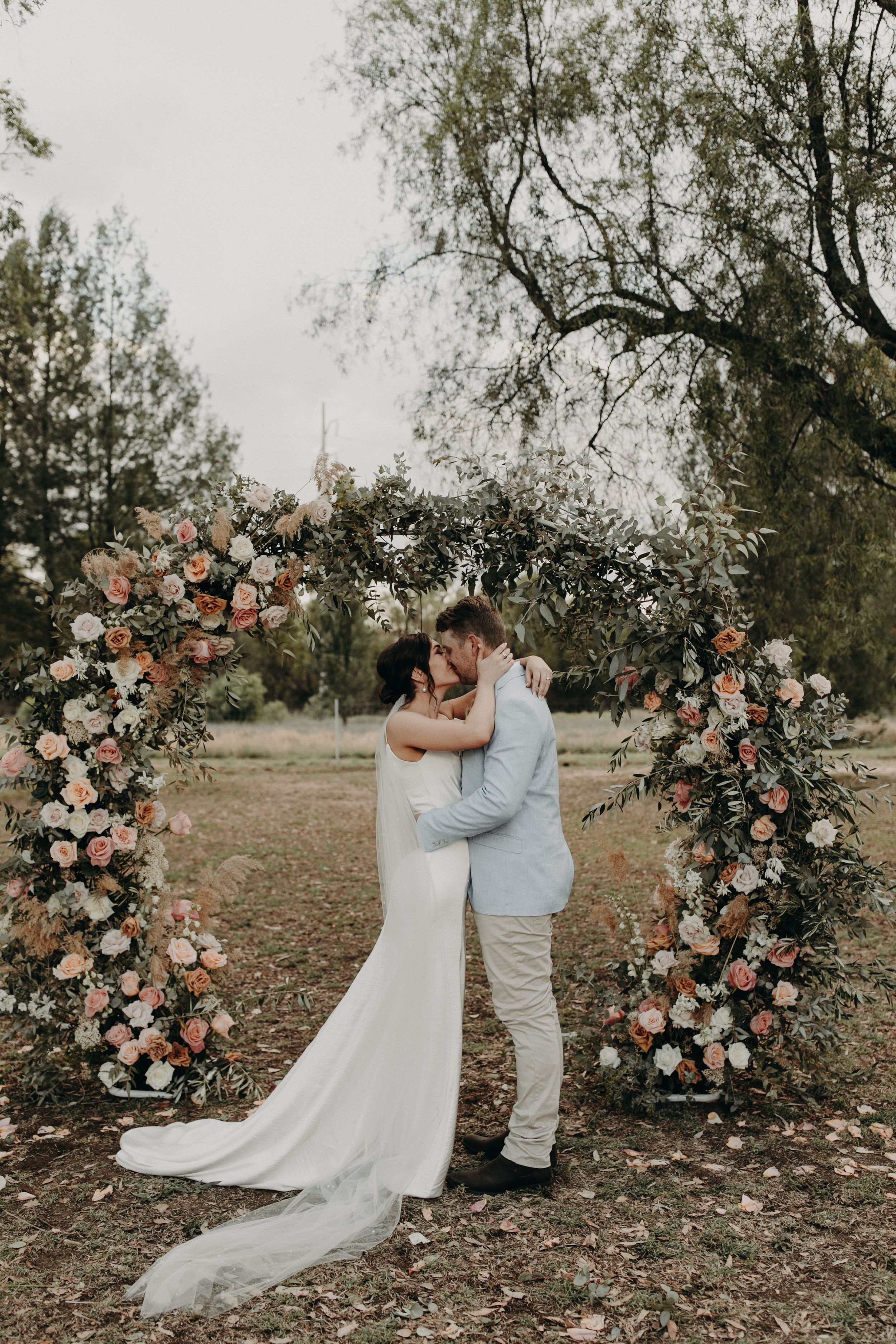 Wedding couple poses by floral arch with peach and white roses in outdoor setting.