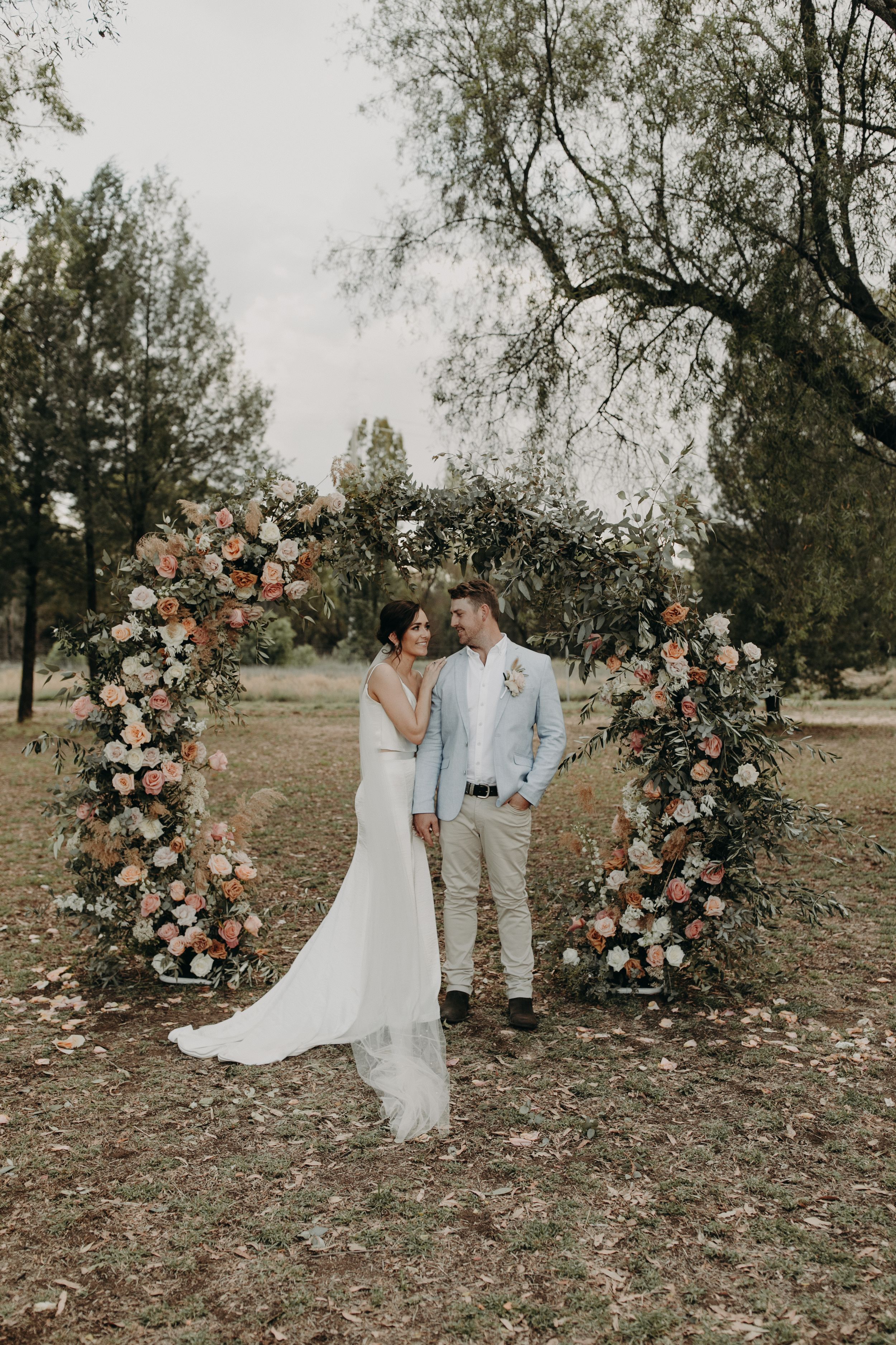 Romantic wedding portraits by floral arch in rustic outdoor setting.