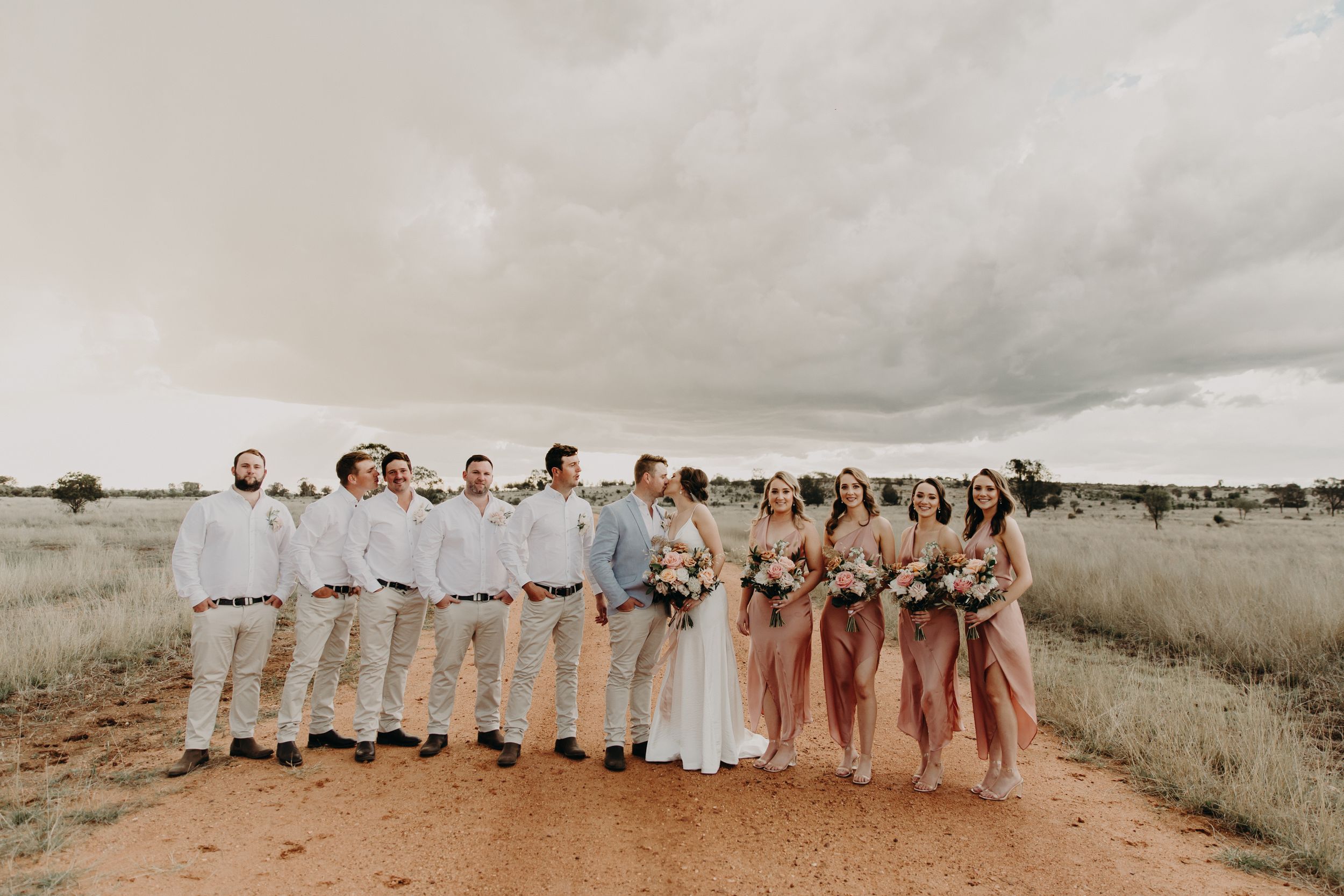 Wedding party in white and blush attire poses together in rustic outdoor setting.