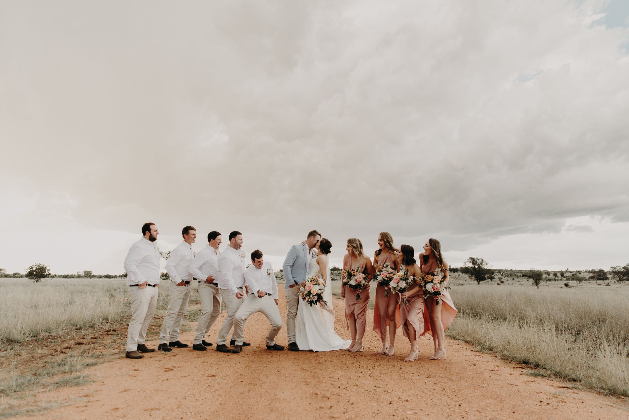 Wedding party lined up in white and blush outfits against cloudy desert landscape.
