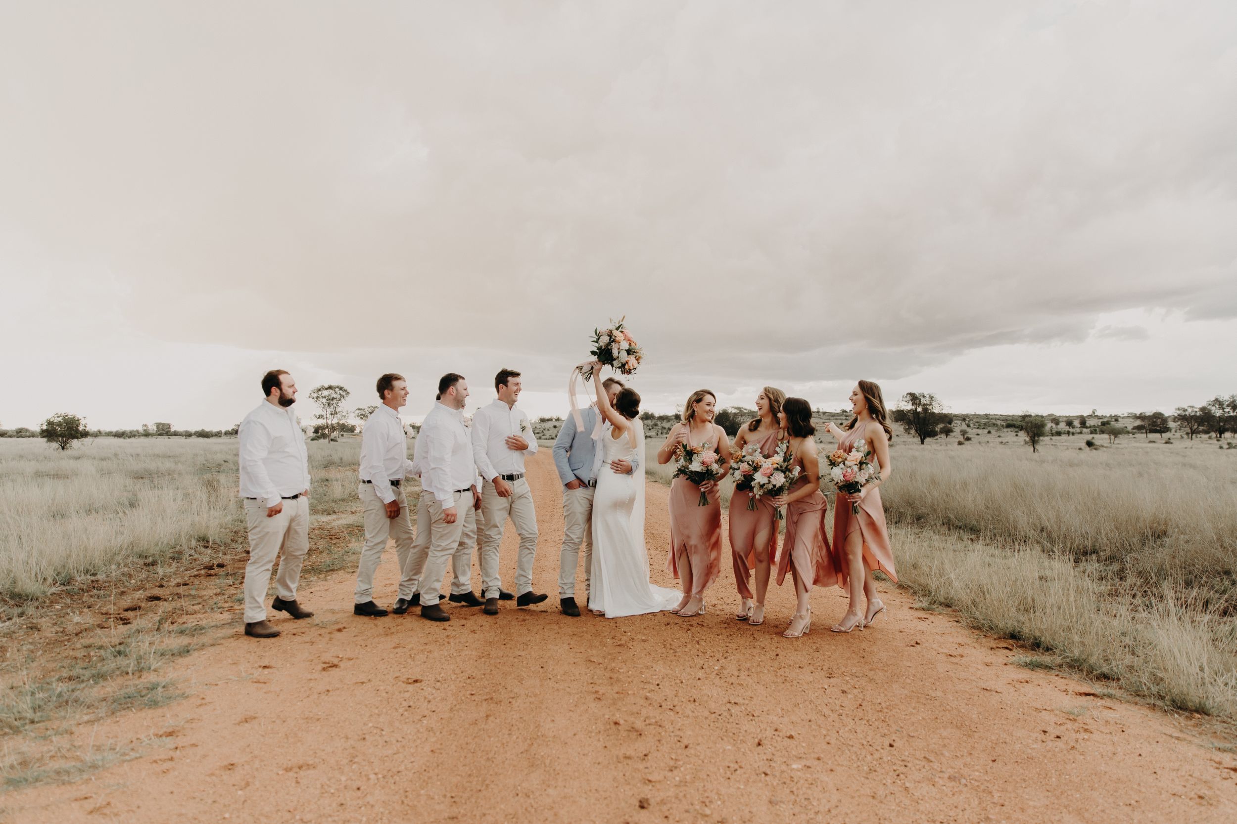 Wedding party arranged in casual poses across desert landscape with cloudy sky.