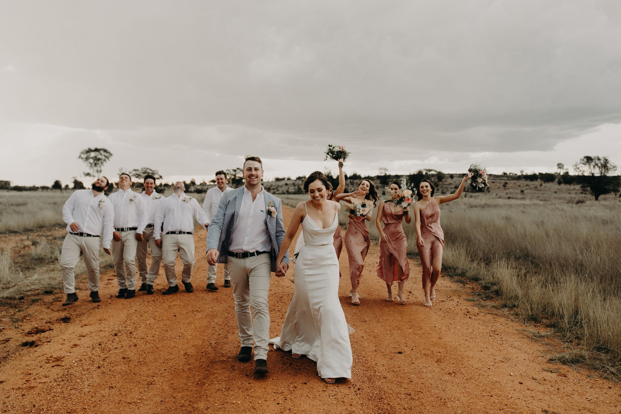 Wedding party walks along a rustic dirt path under moody skies at a countryside venue.