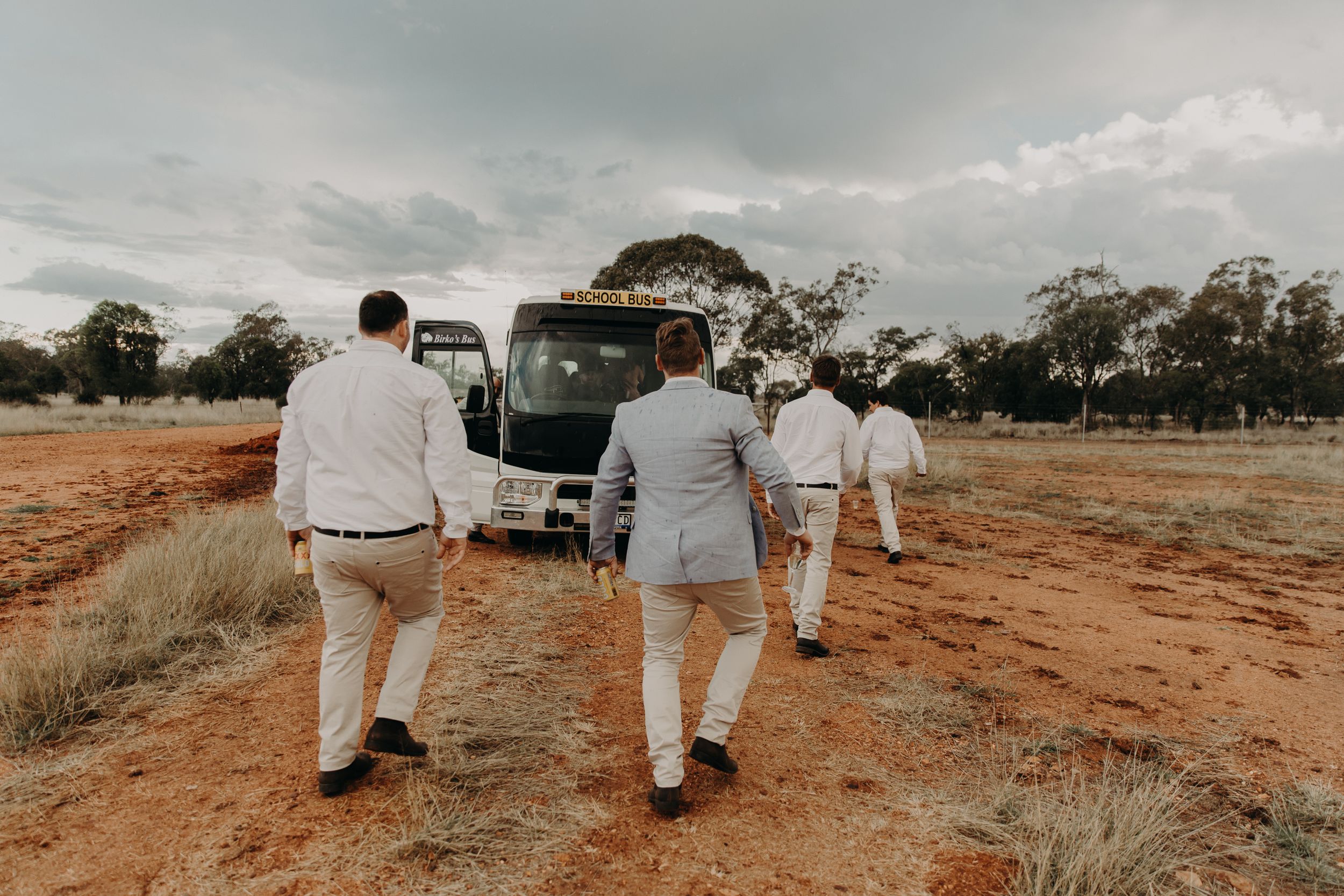 Wedding guests gather on a red dirt path surrounded by dry grassland and overcast sky.