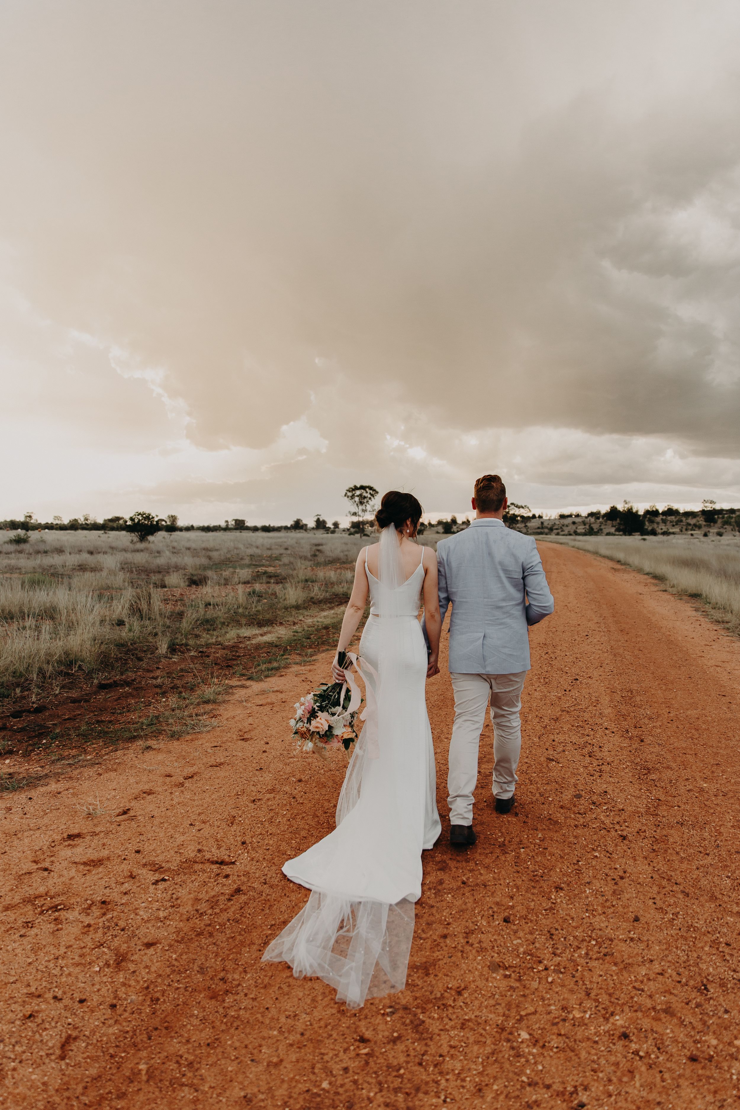 Couple walks hand in hand down a red dirt road with stormy skies in the background.