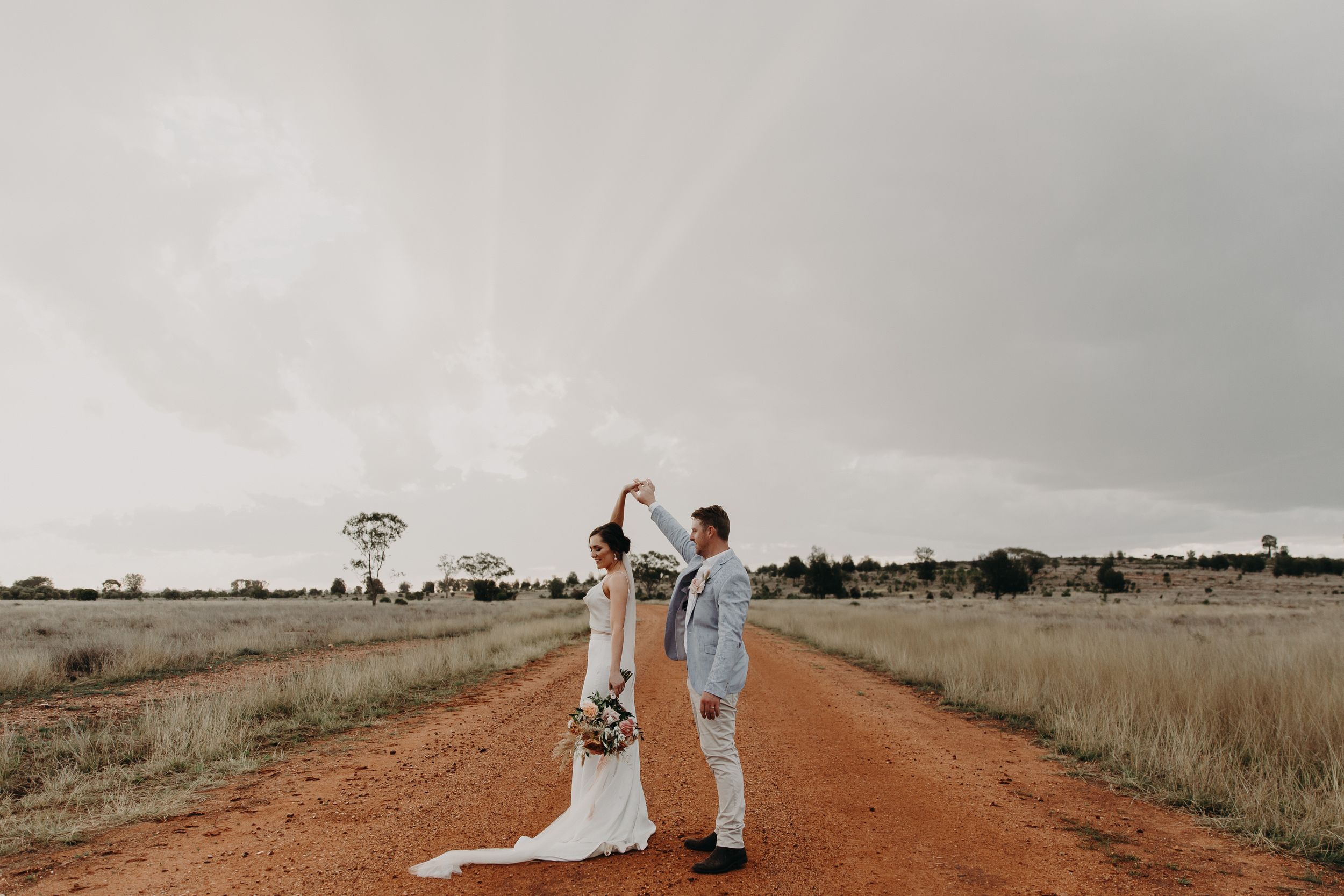 A couple dances on a remote red dirt road with storm clouds and distant trees.