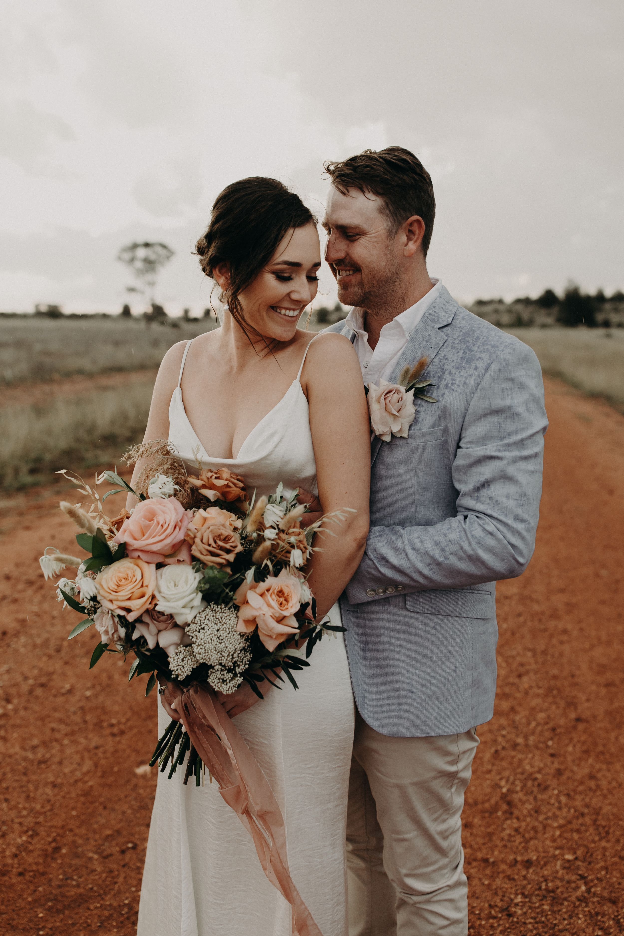A couple poses with a peach and white floral bouquet on a rustic outback road.