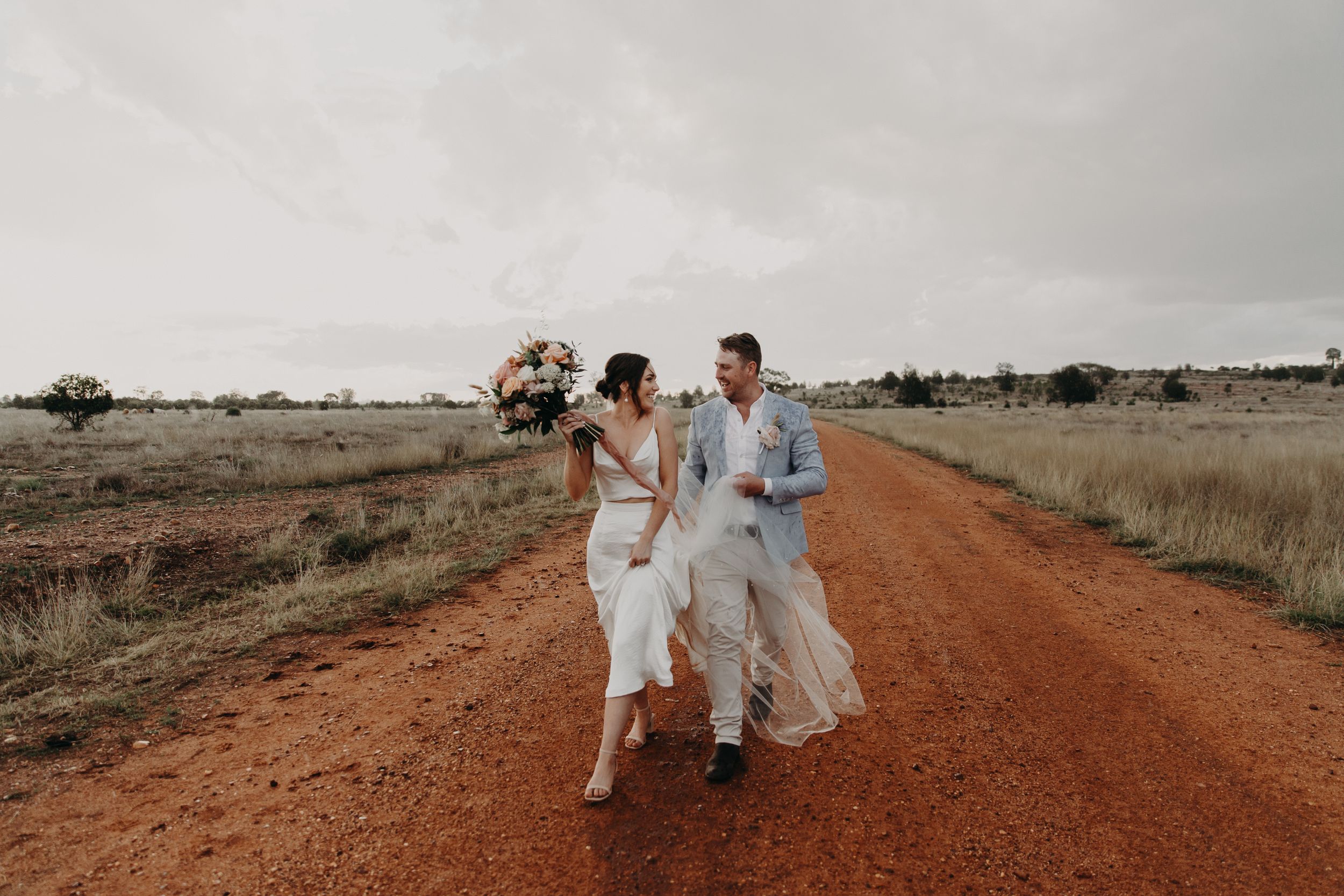 A couple walks along a rustic red dirt road in the Australian outback during a wedding photoshoot.
