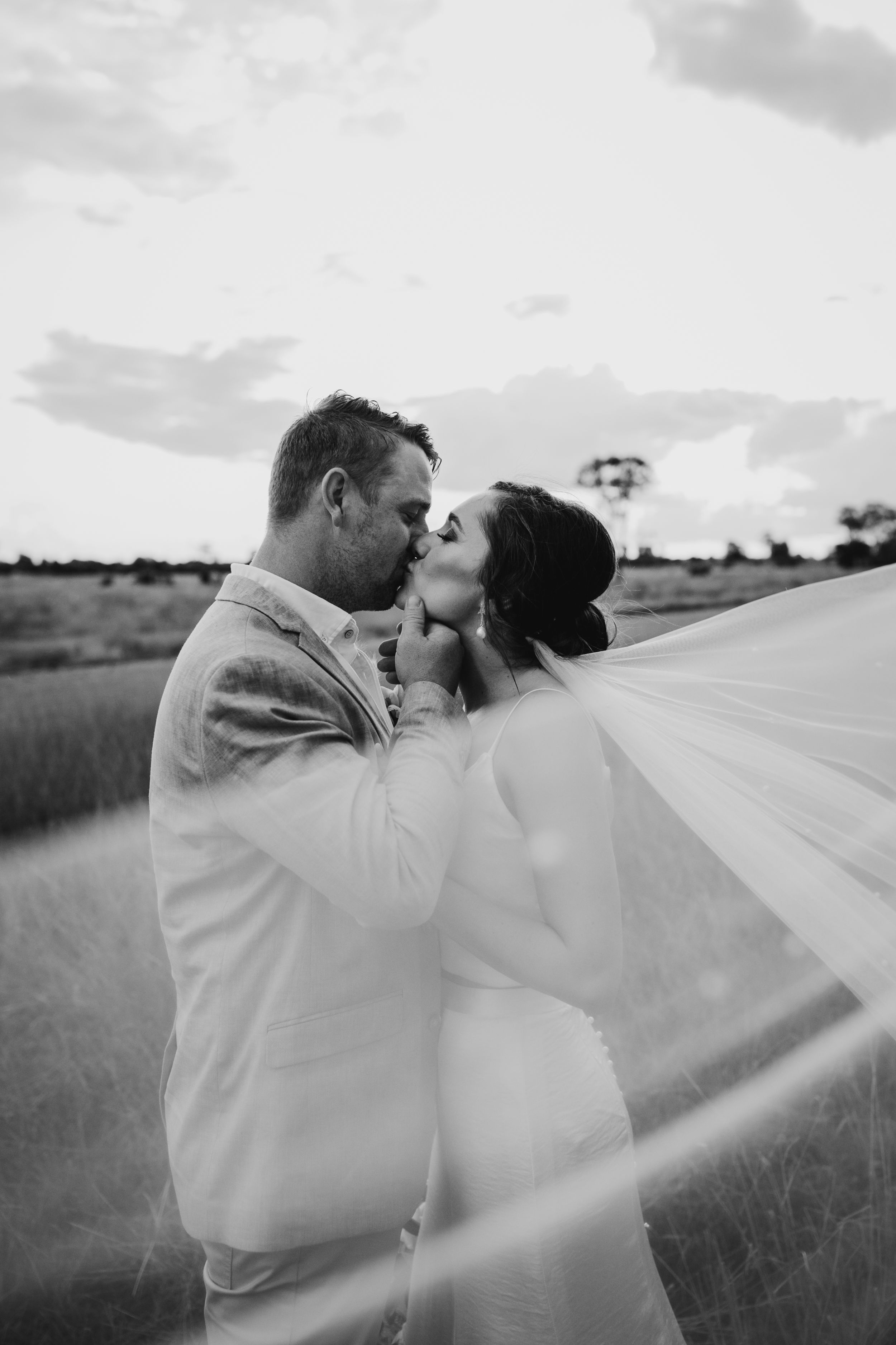 A black and white portrait of newlyweds with flowing veil in rural setting.