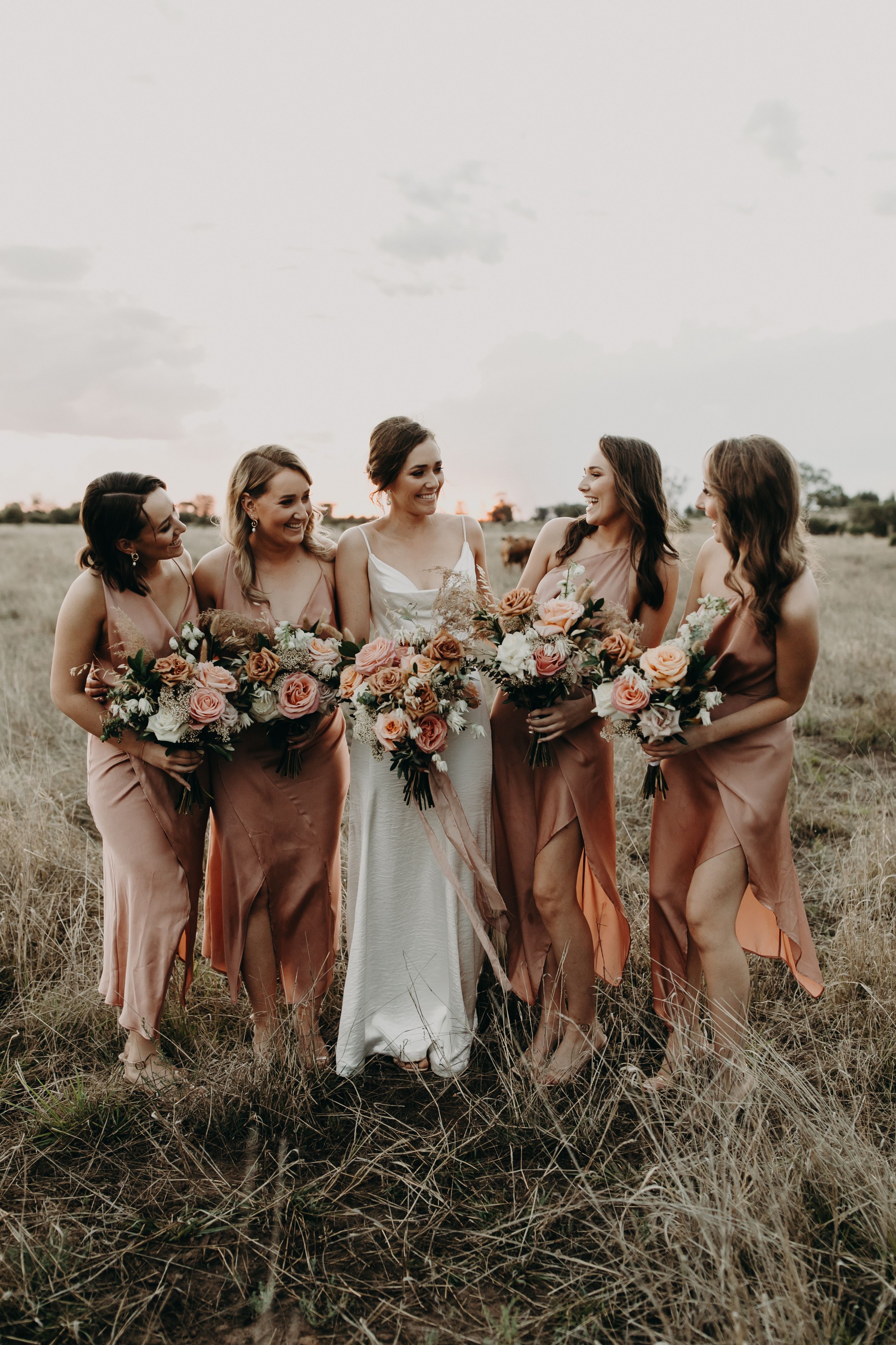 Wedding party of bridesmaids in mauve dresses share candid moment with bride.