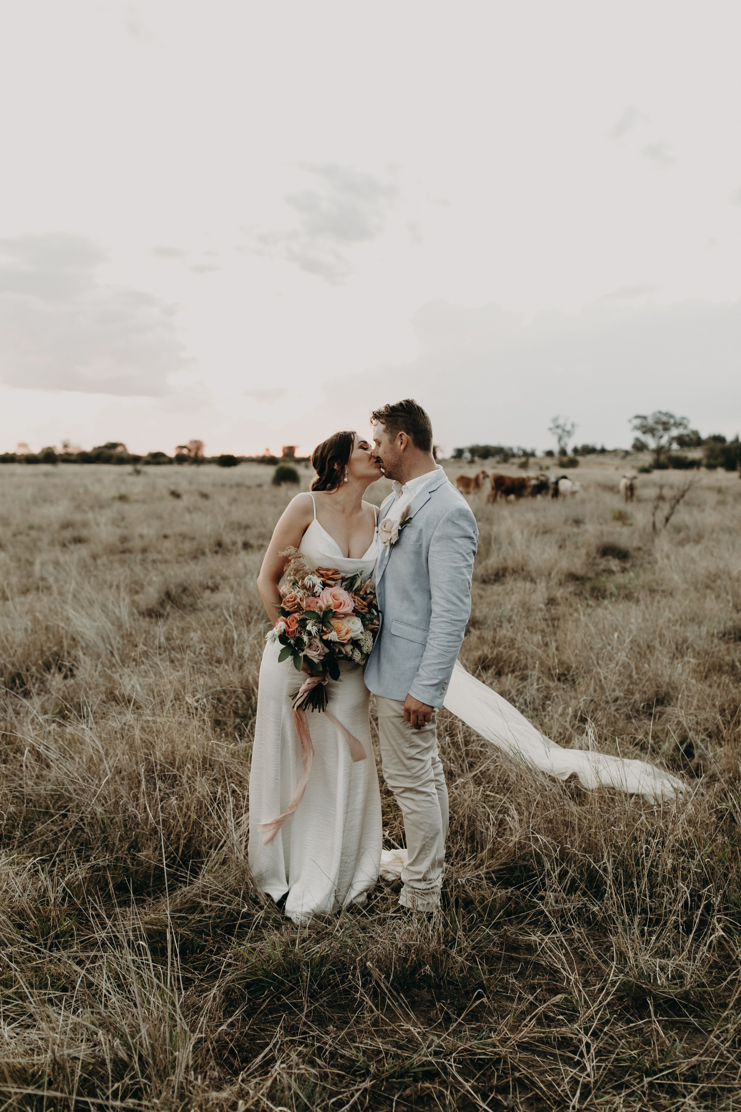 Newlyweds share intimate moment in rustic field with dramatic sky backdrop.