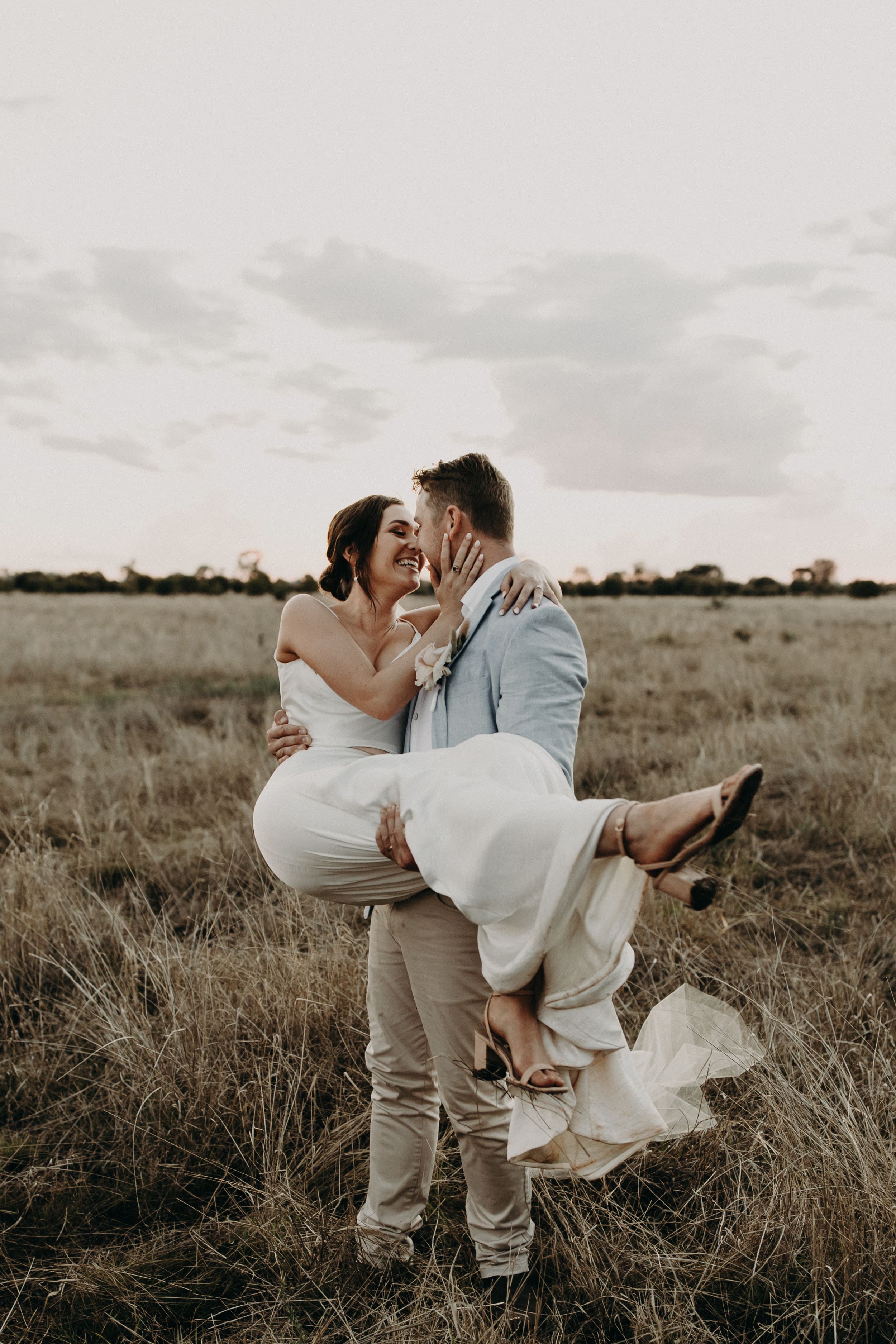 A couple shares an intimate moment in a field during golden hour as her white dress flows in the wind.