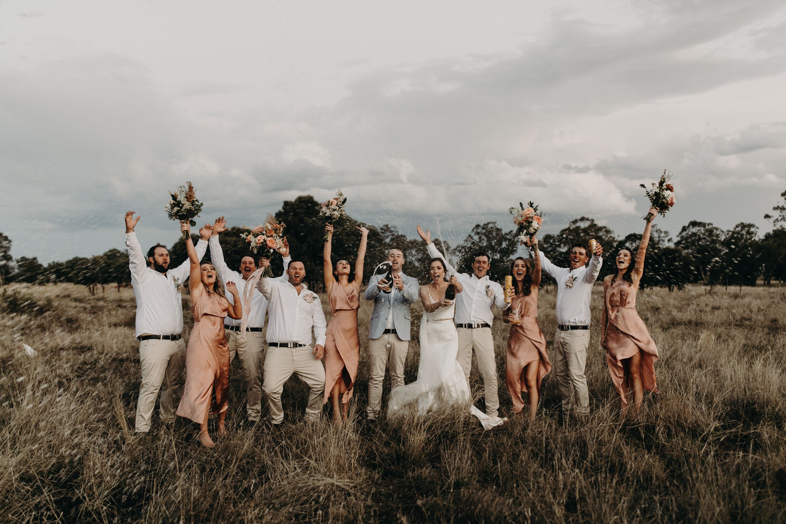Wedding party jumps joyfully for a group photo in an outdoor field setting.