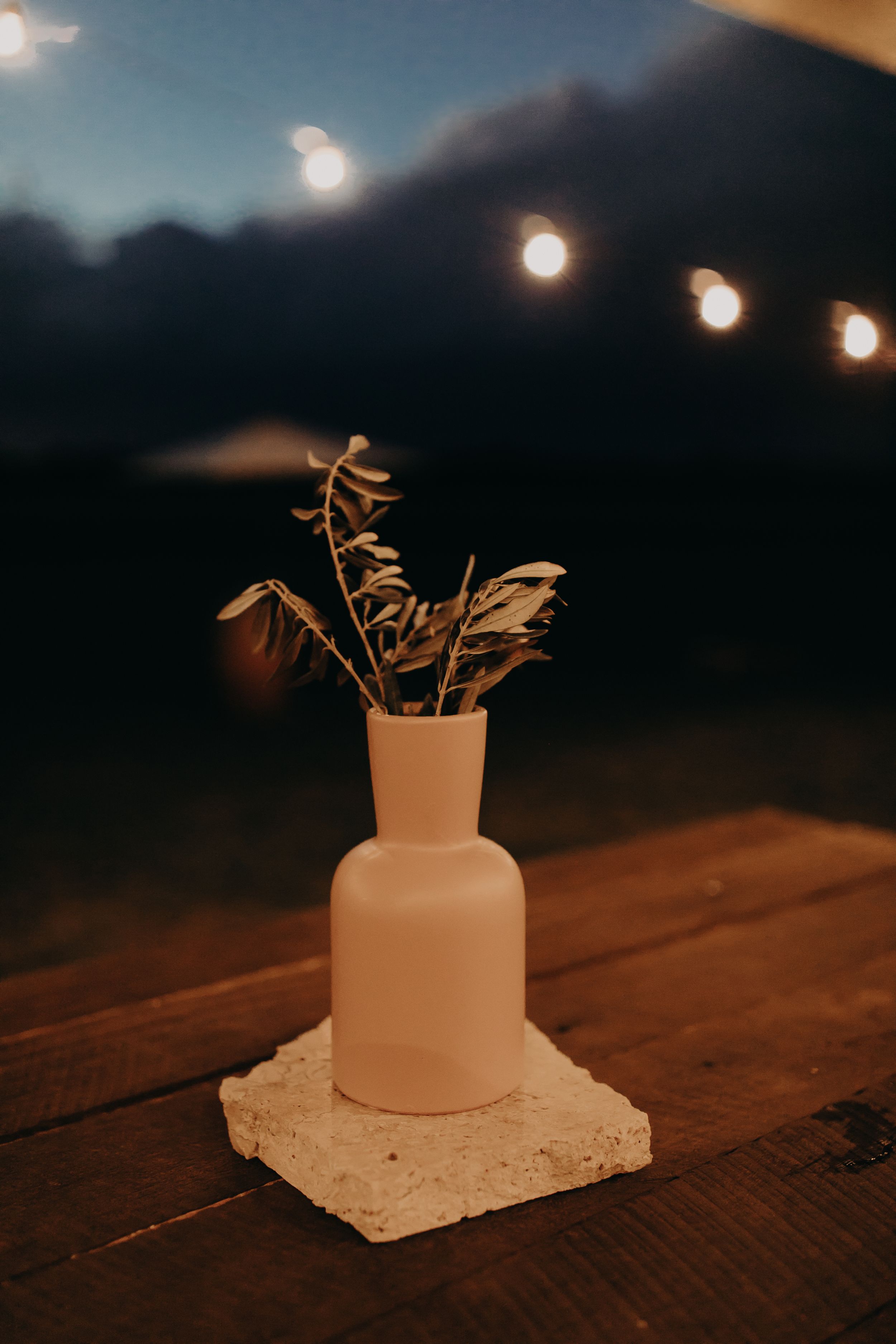 Small white vase with delicate dried flowers sits on a wooden surface under string lights at dusk.