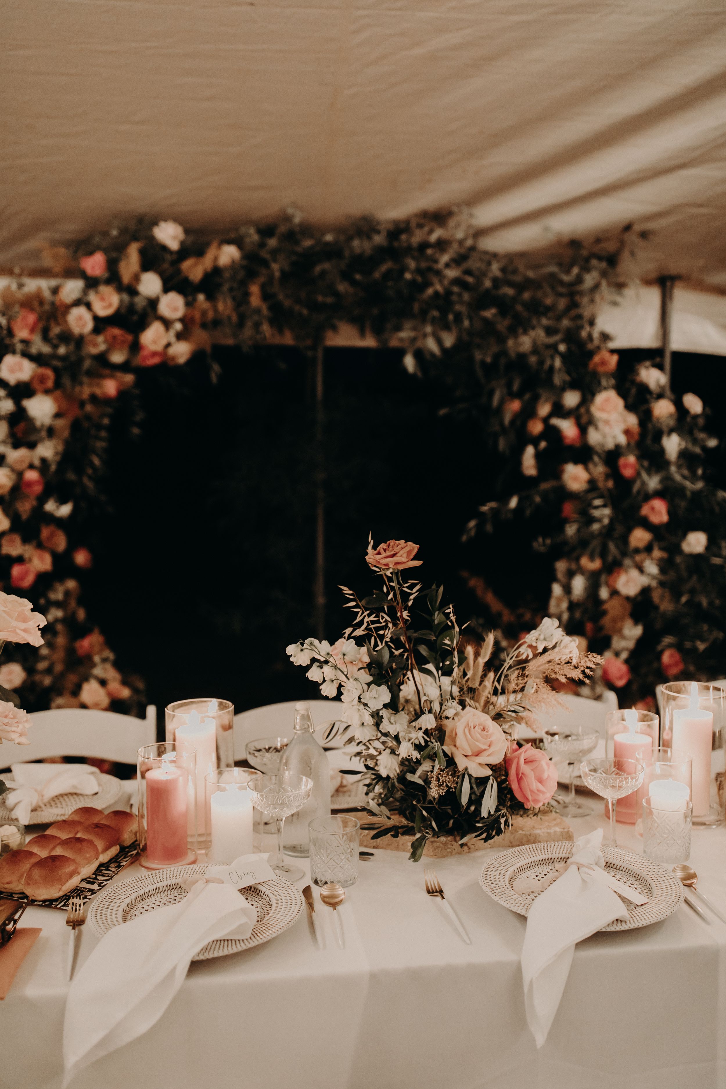 Wedding table decorated with pink roses, candles and place settings under tent lighting.
