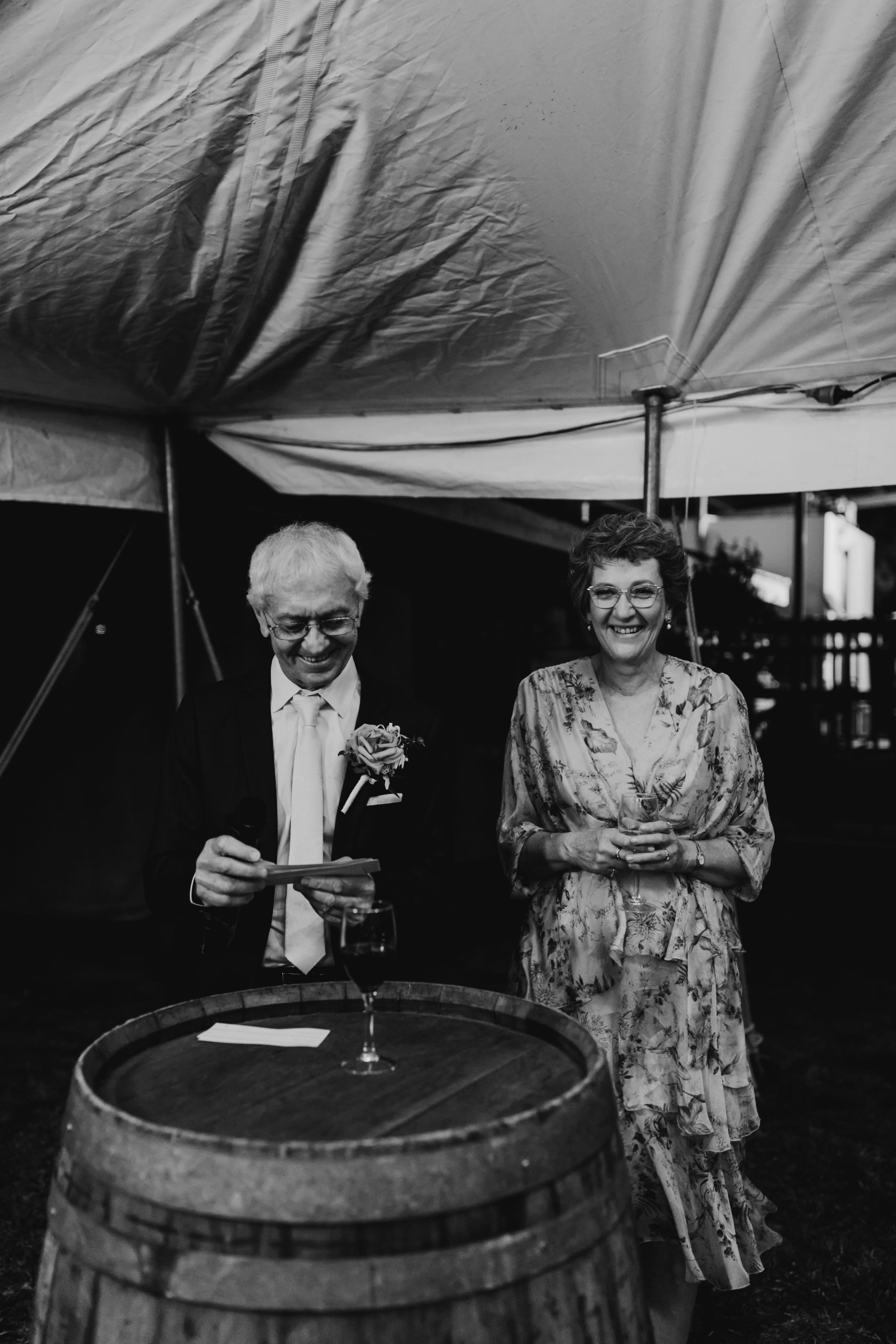Wedding guests enjoying drinks around wine barrels under a tent at dusk.