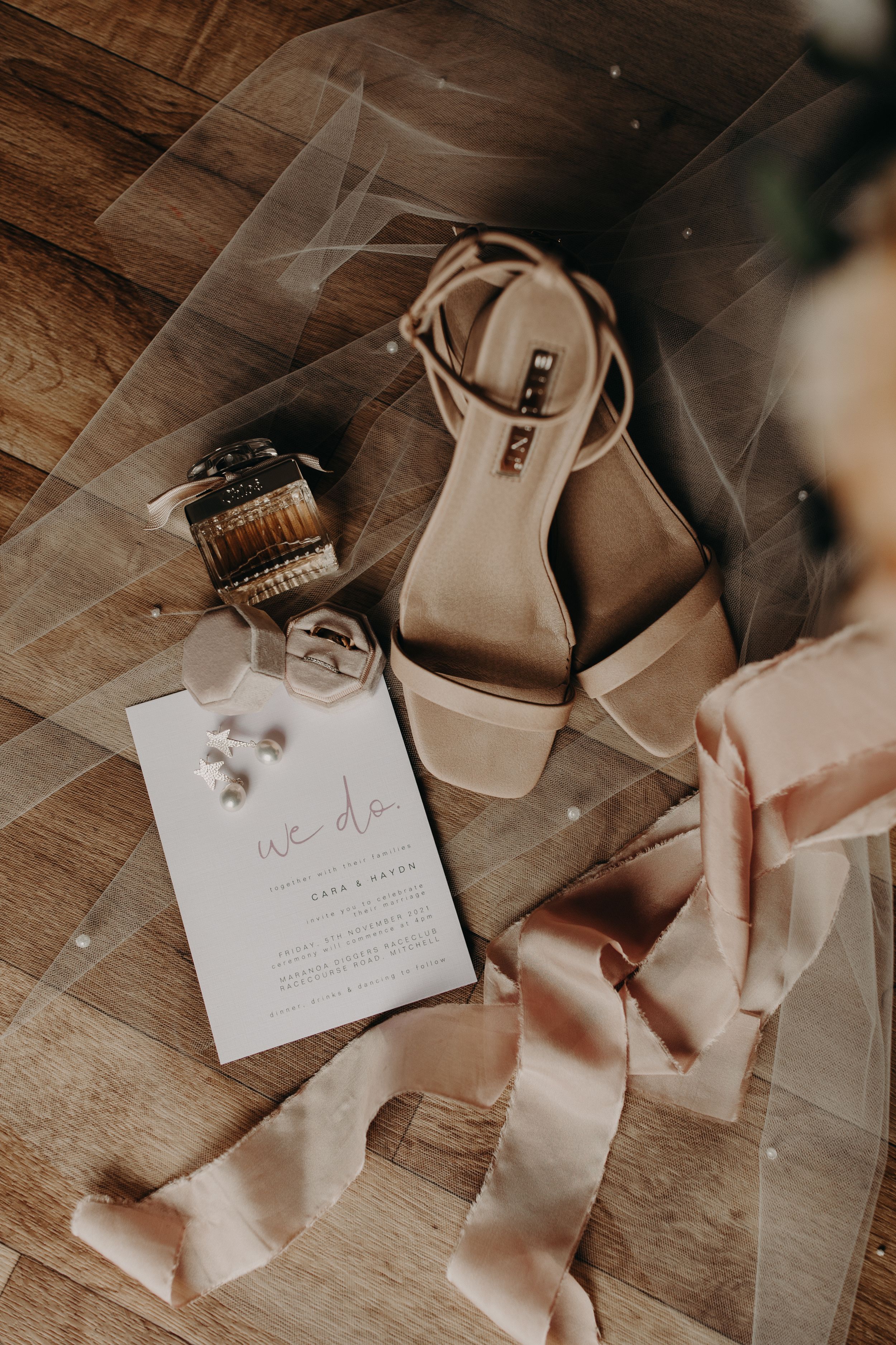 Wedding accessories including invitation, shoes, and silk ribbon arranged on wooden floor.