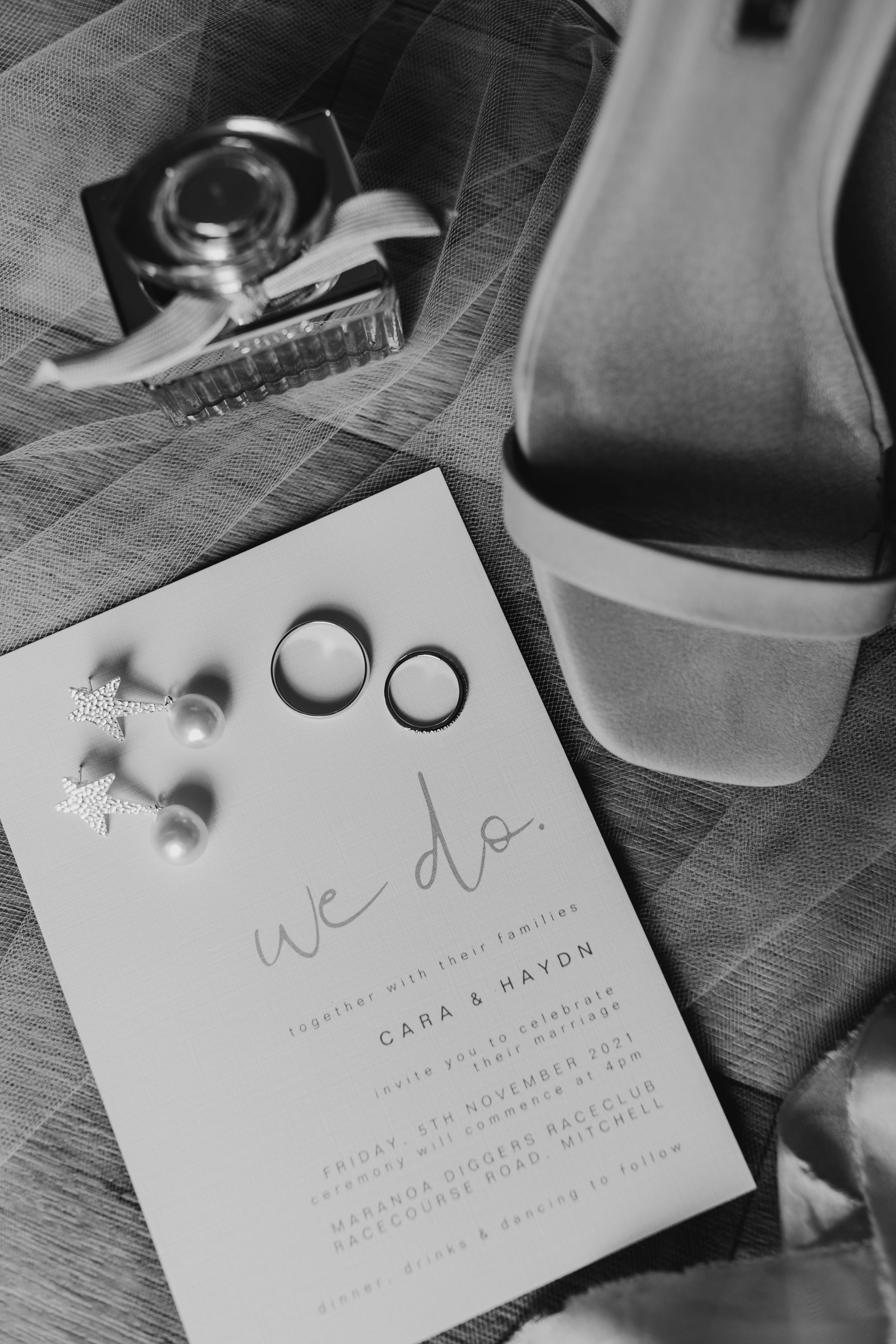 Wedding invitation and rings displayed on dark wooden surface in black and white.