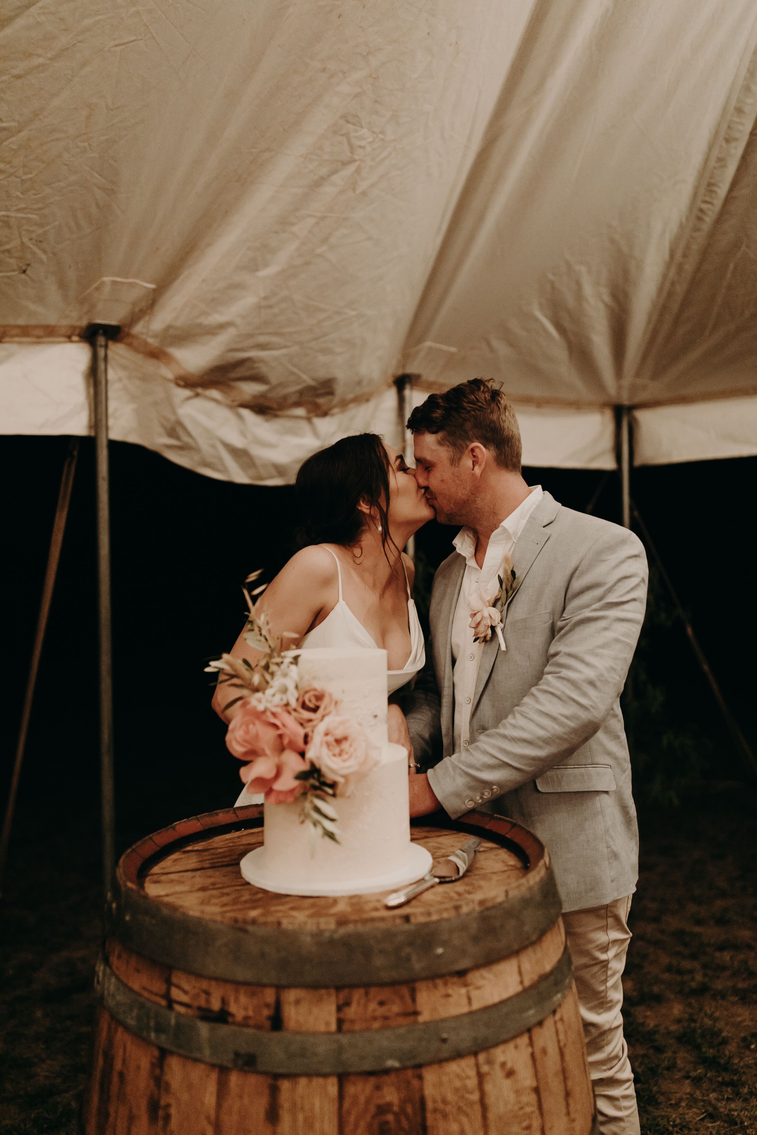 Newlyweds share an intimate moment while cutting their wedding cake on a rustic wine barrel at an outdoor reception.