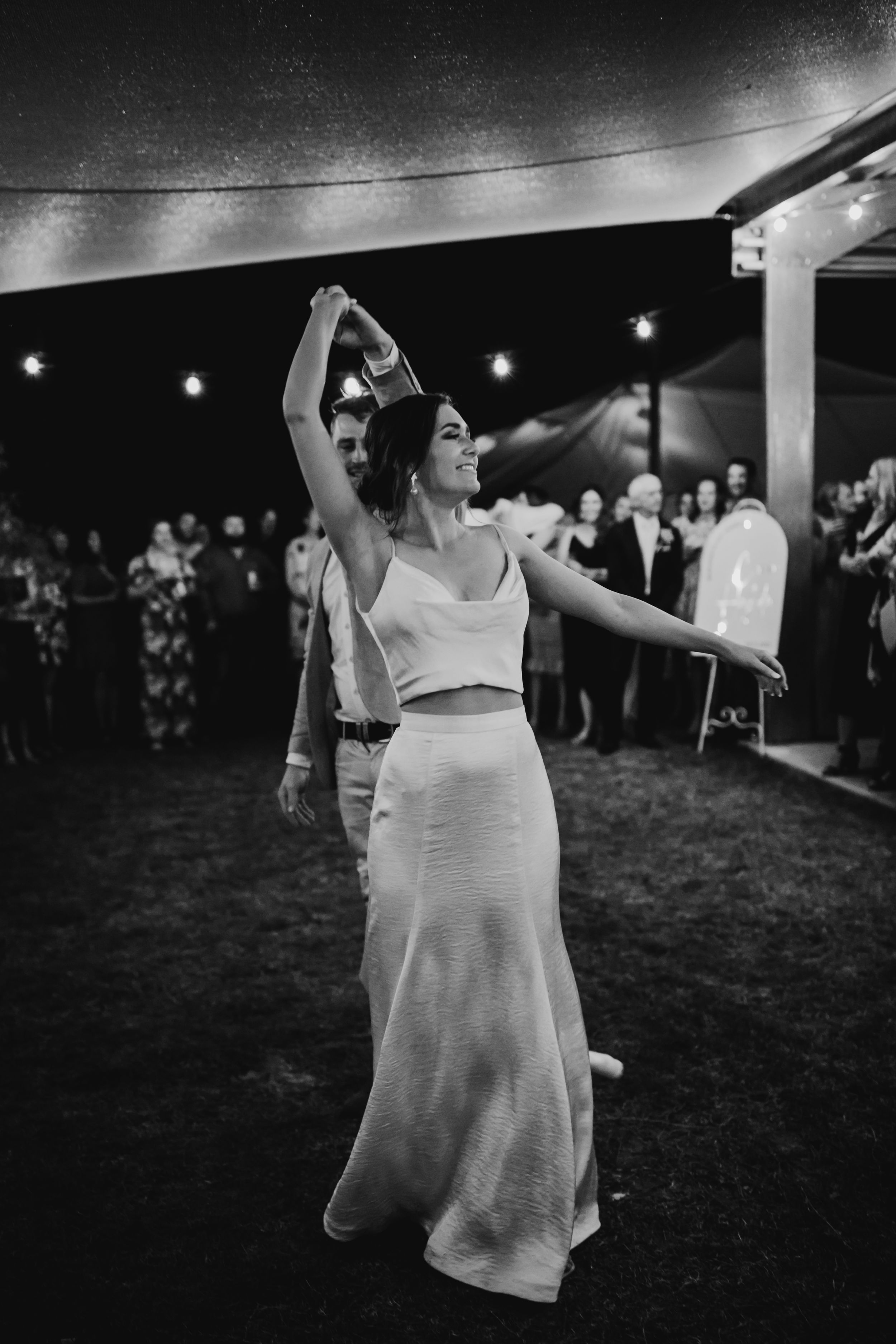 Wedding guests dance and celebrate under string lights at an outdoor evening reception.